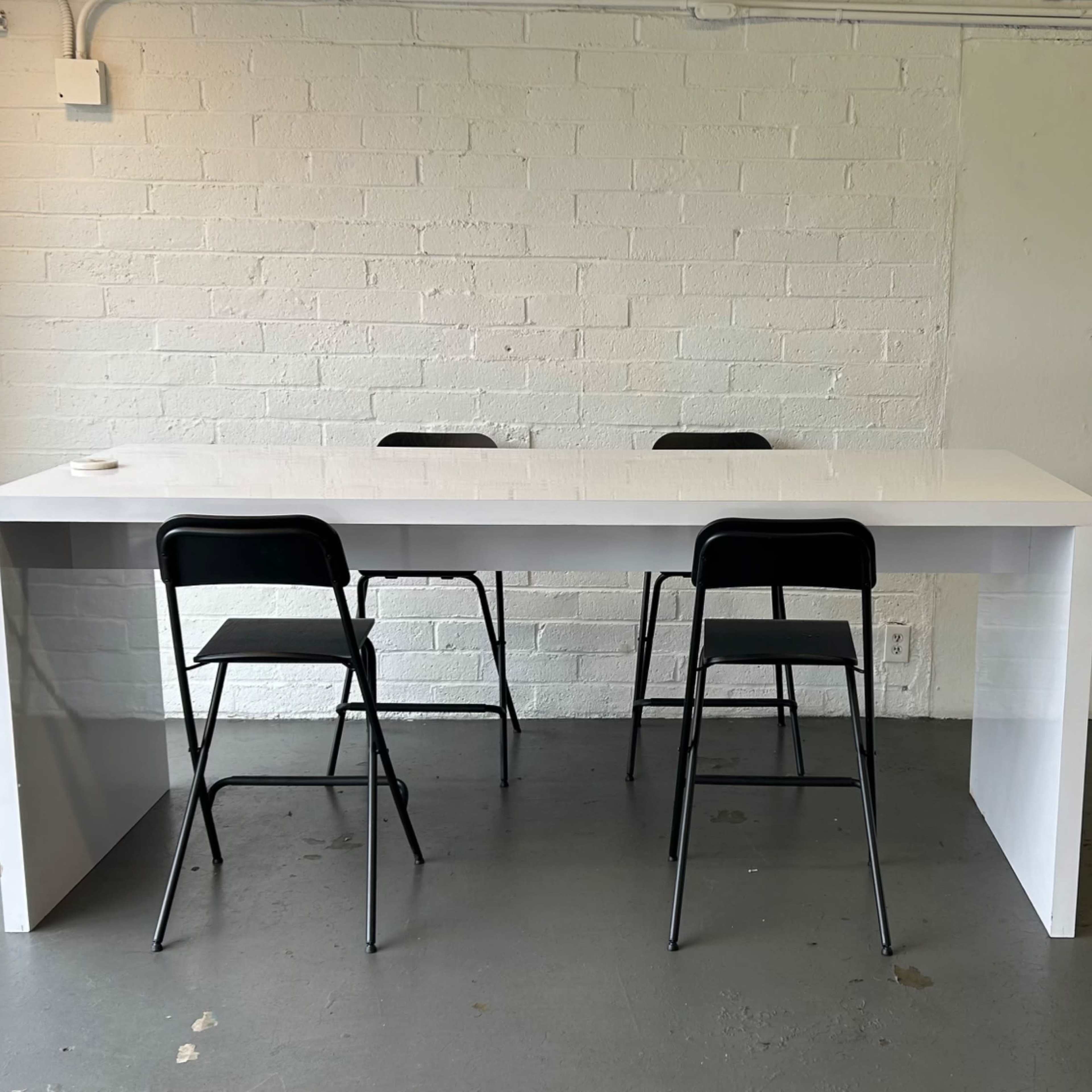 A white table with four black chairs is positioned in a room with a textured white brick wall and gray flooring.