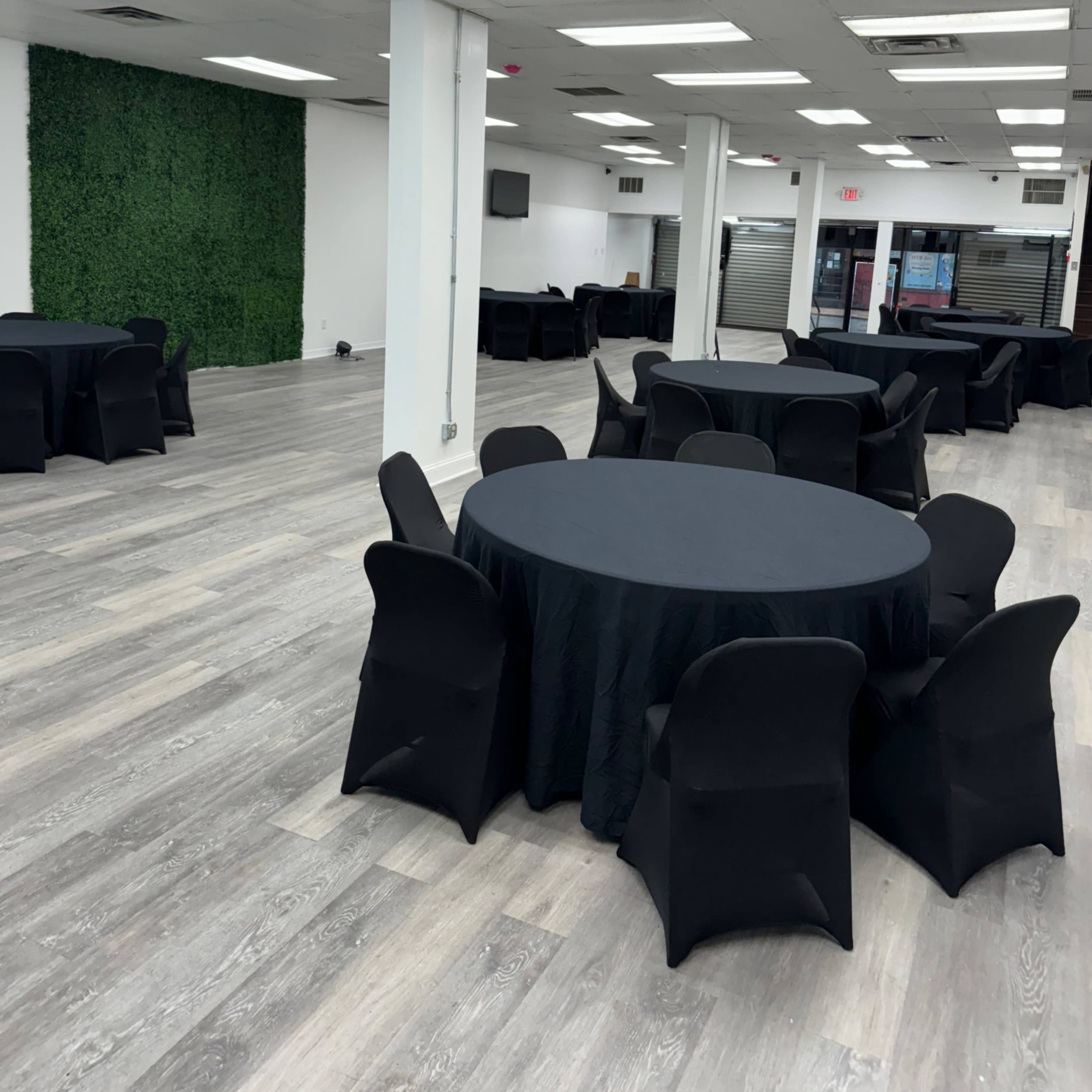A spacious event room with several round tables covered in black tablecloths and a decorative green wall in the background.