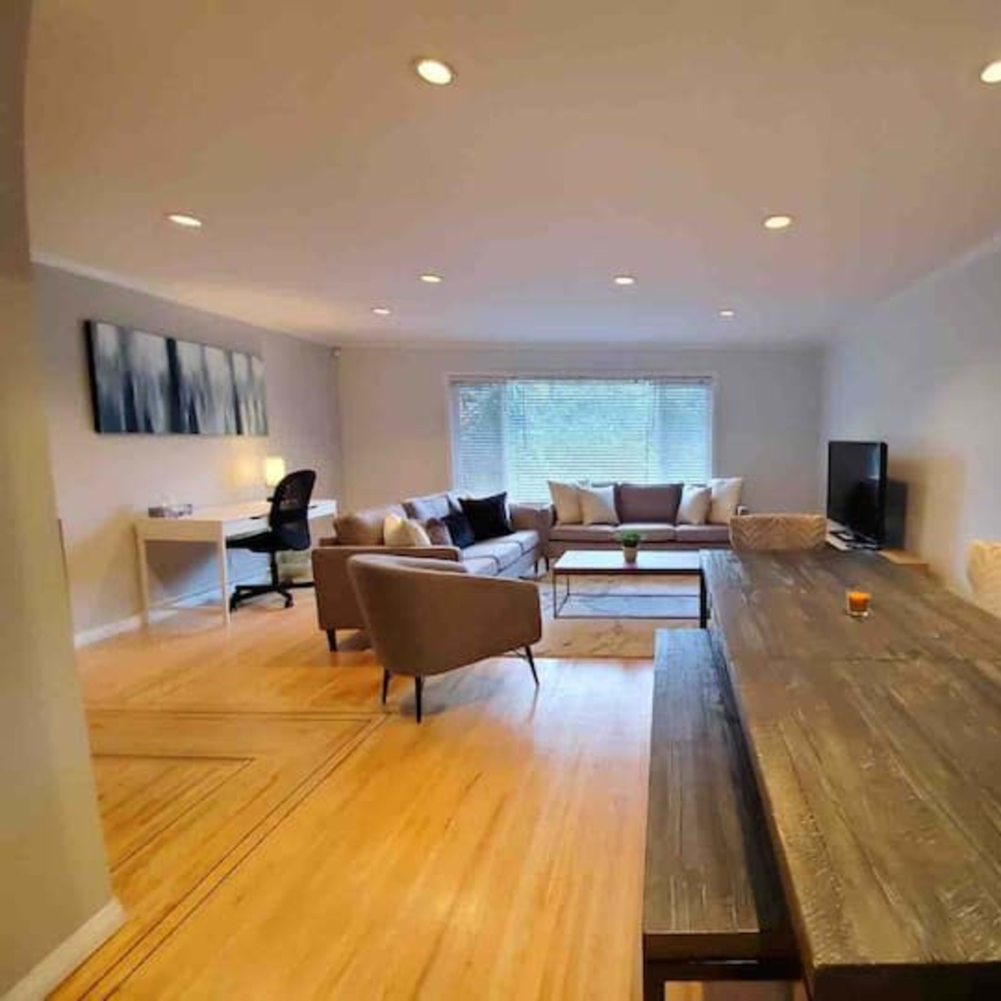 A modern living room with a wooden dining table, a sofa arrangement, a television, and a desk with a chair, all illuminated by recessed lighting.