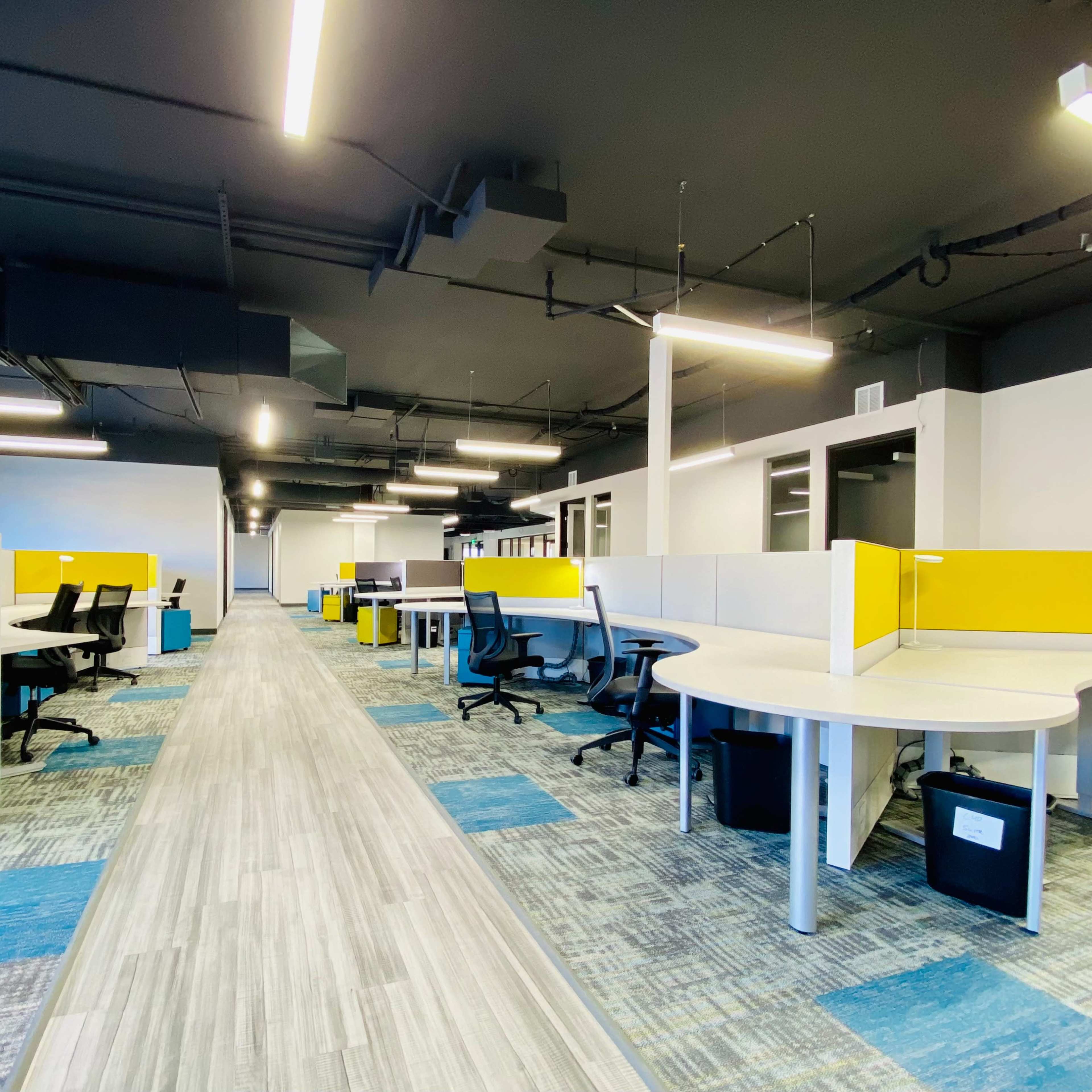 A modern office space with several workstations featuring yellow dividers, gray flooring, and overhead lighting.