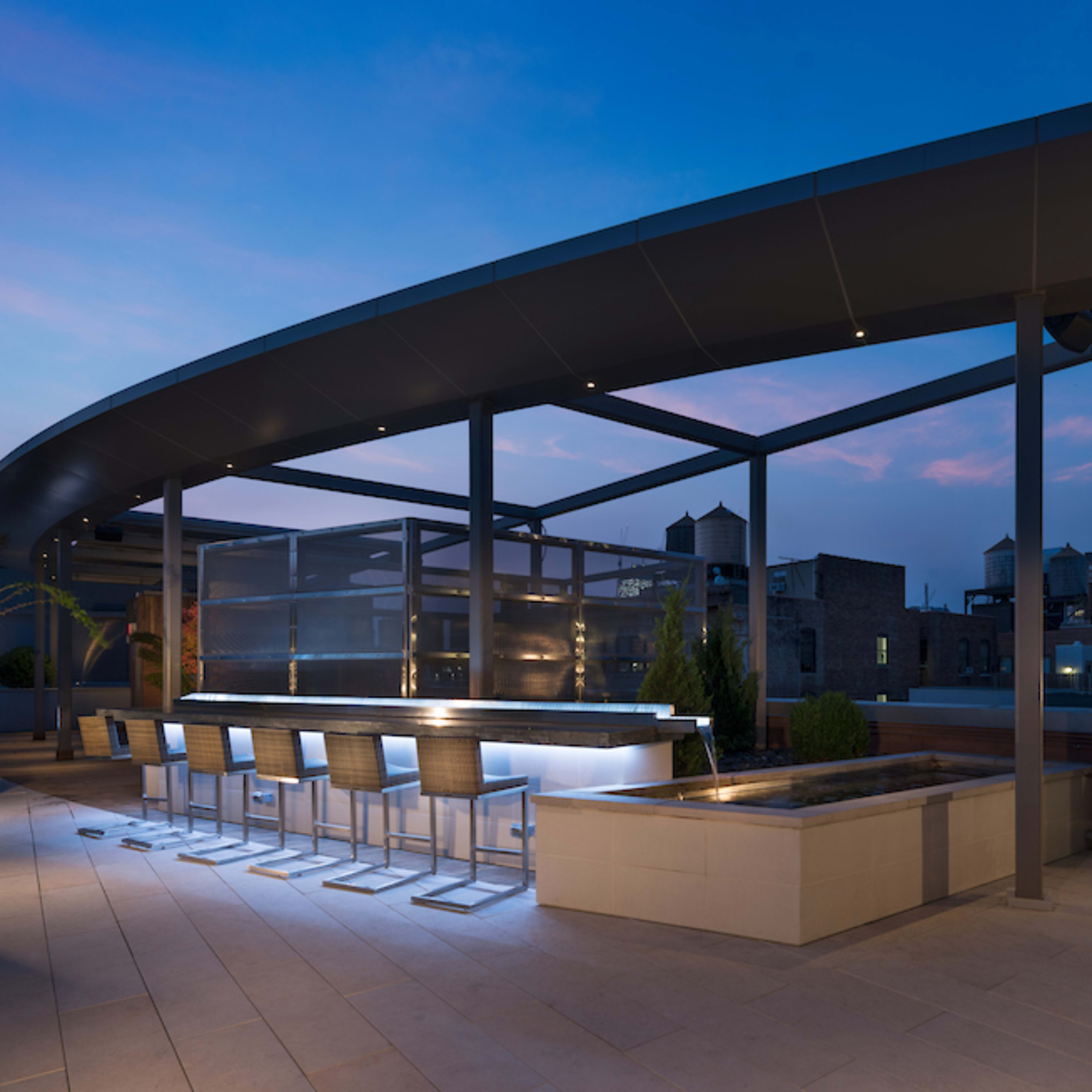 A modern rooftop terrace features a sleek bar area, planters, and a view of a city skyline at dusk.