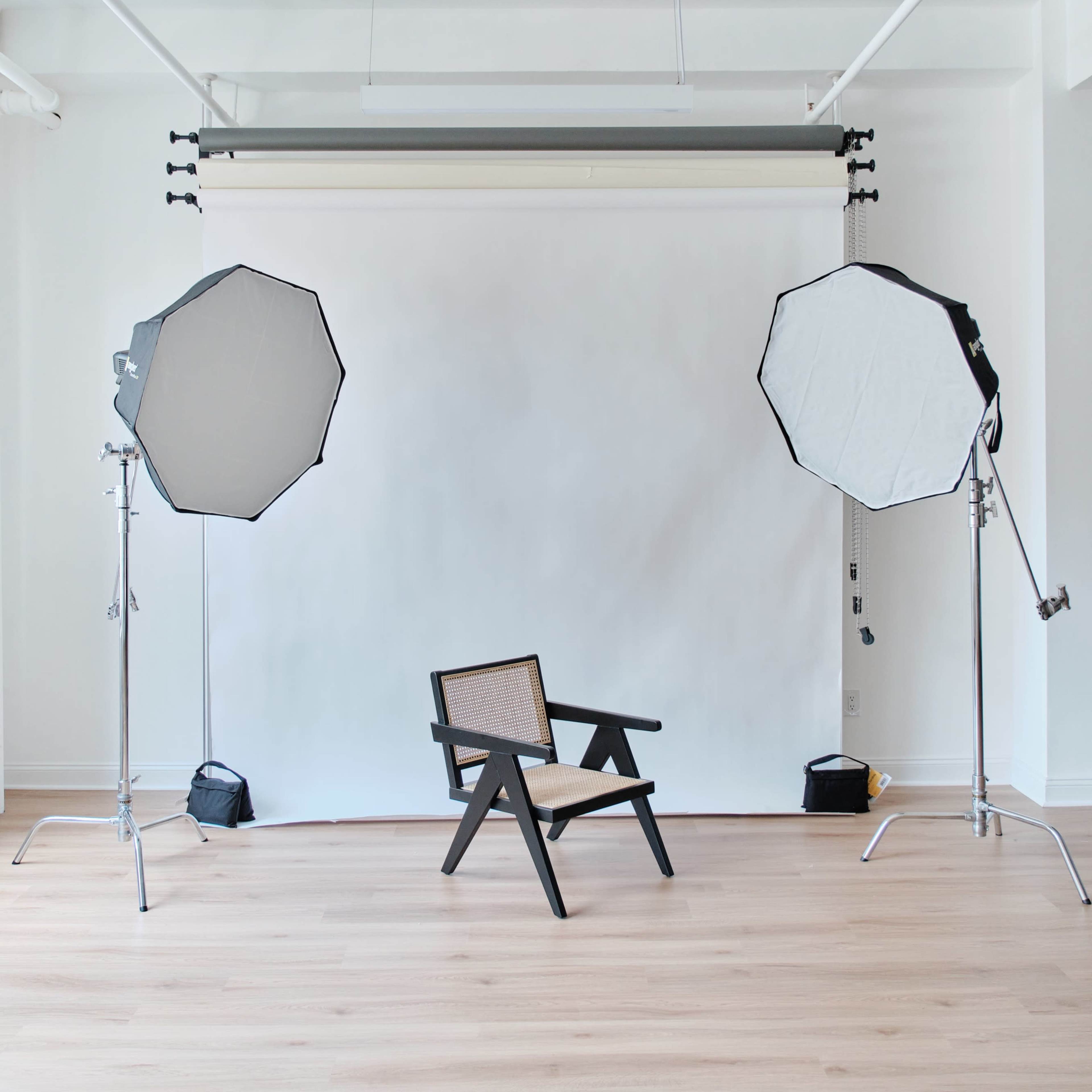 A minimalist photography studio features a black chair positioned in front of a white backdrop, flanked by two large softbox lights.