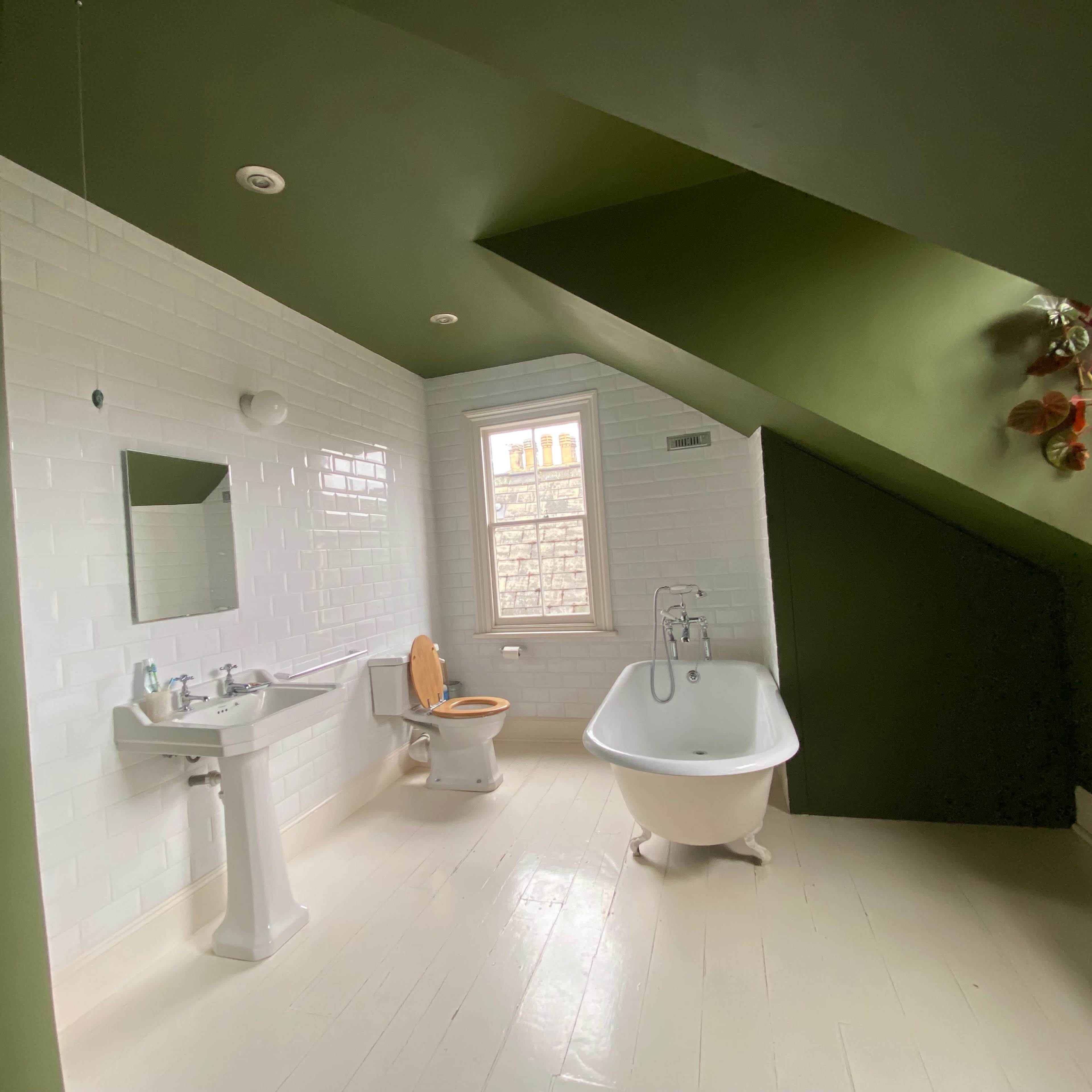 A bathroom with white tiled walls, a green sloped ceiling, a freestanding bathtub, a sink, a toilet, and a window.