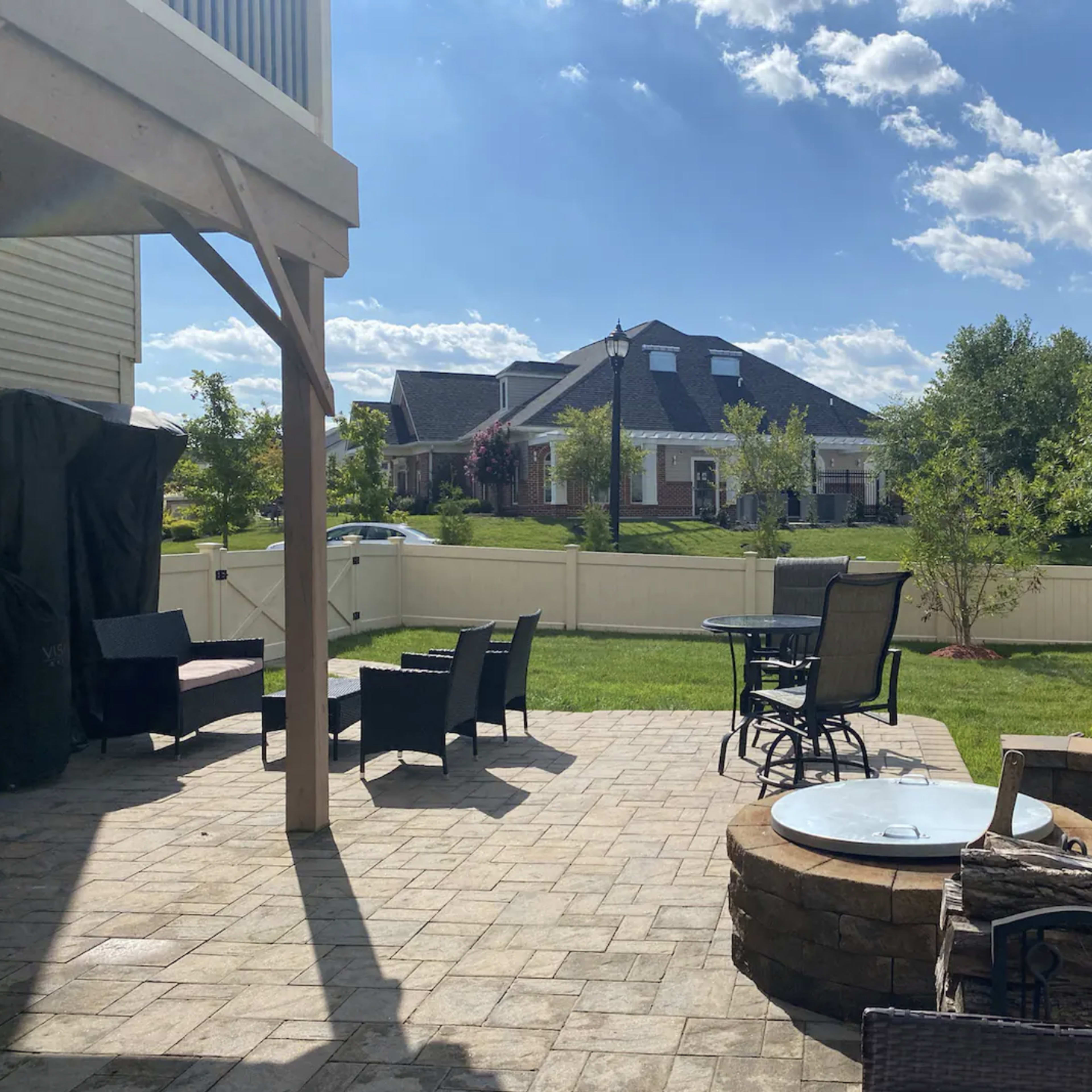 A patio area with seating and a small fire pit, surrounded by a well-maintained lawn and a house in the background under a partly cloudy sky.