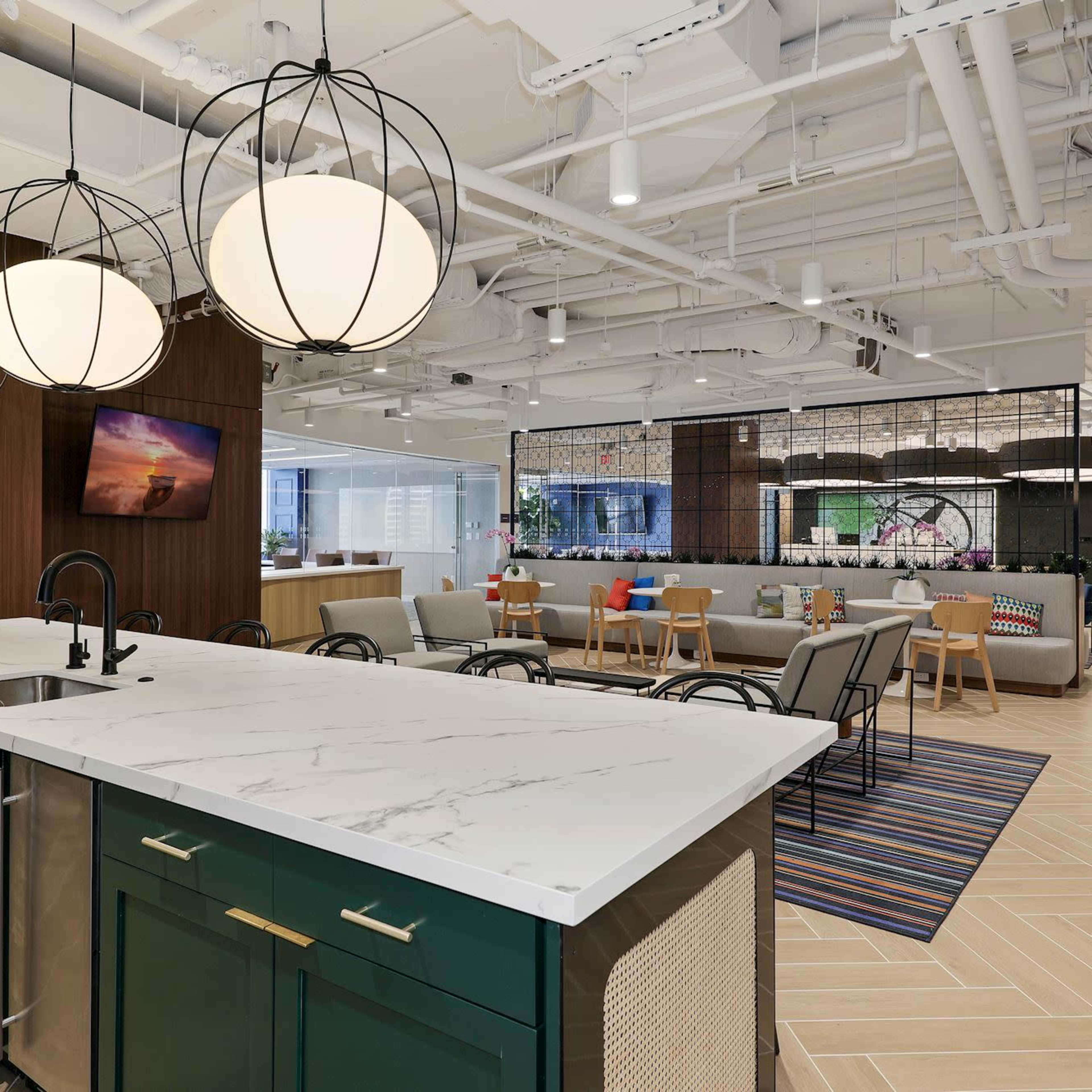 A modern office kitchen with a large island, pendant lighting, a seating area, and a combination of wood and metal furnishings.