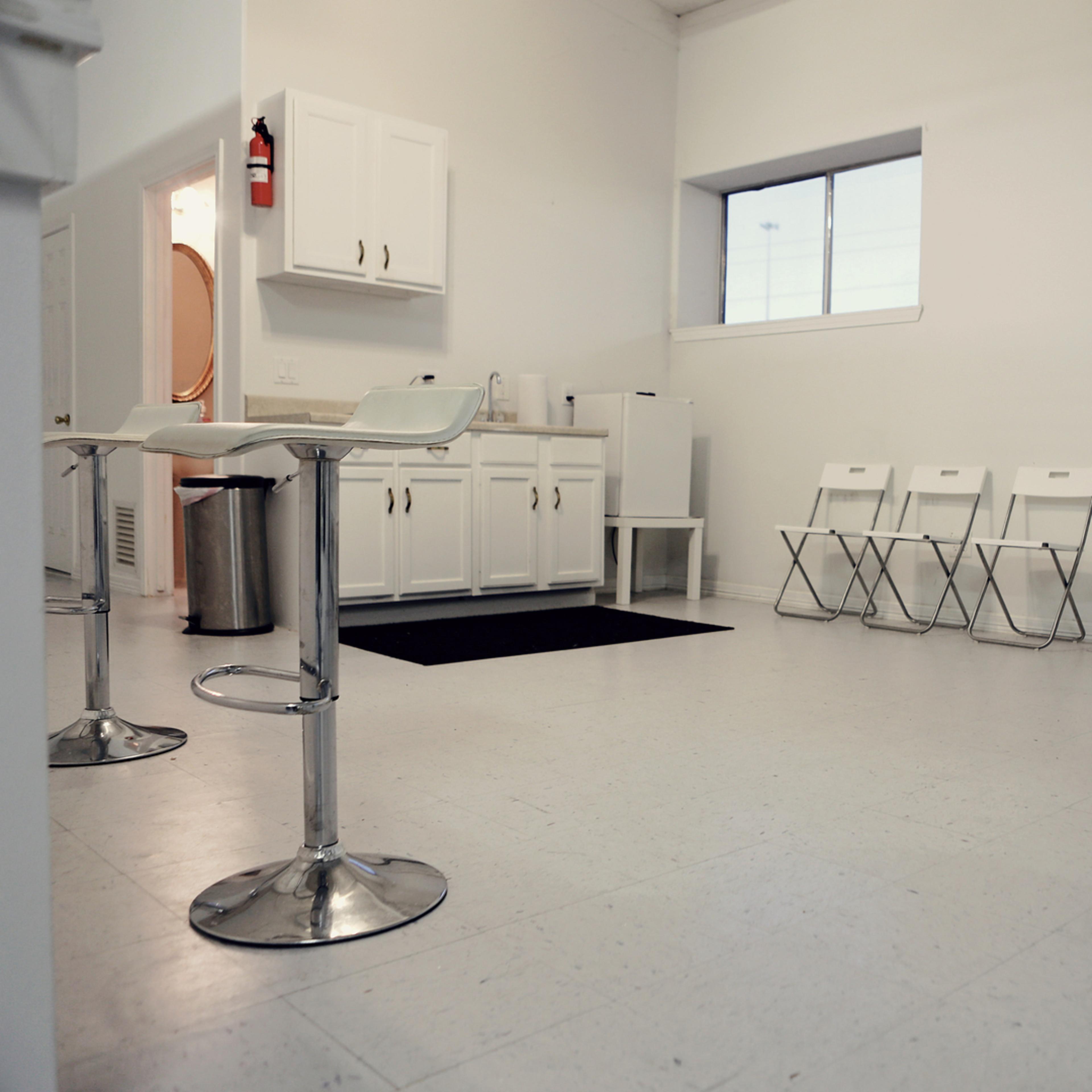 A sparsely furnished waiting area with a bar table, stools, and a row of white folding chairs against a white wall.