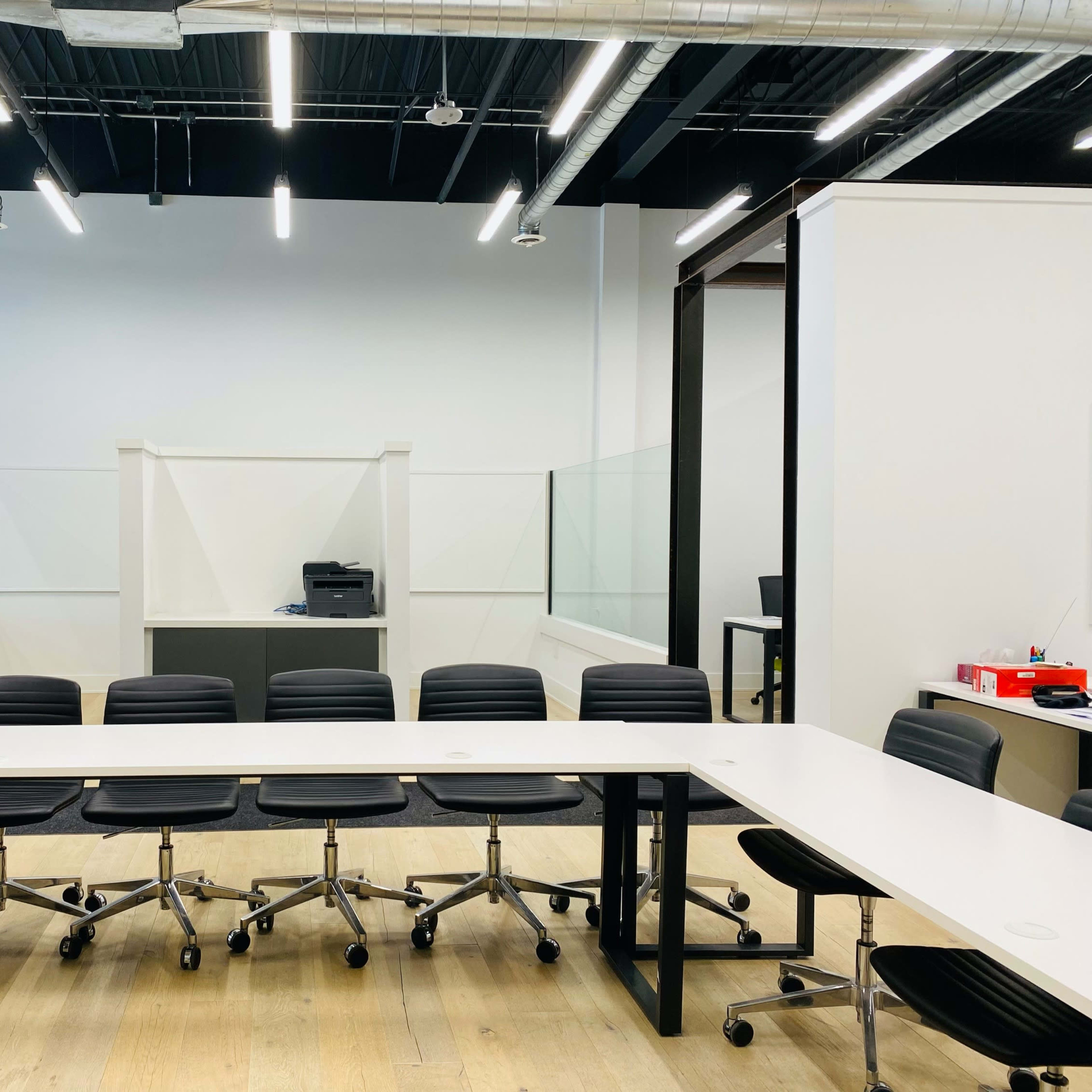 A modern conference room with a long white table and several black rolling chairs arranged around it, flanked by glass partitions and a printer in the background.