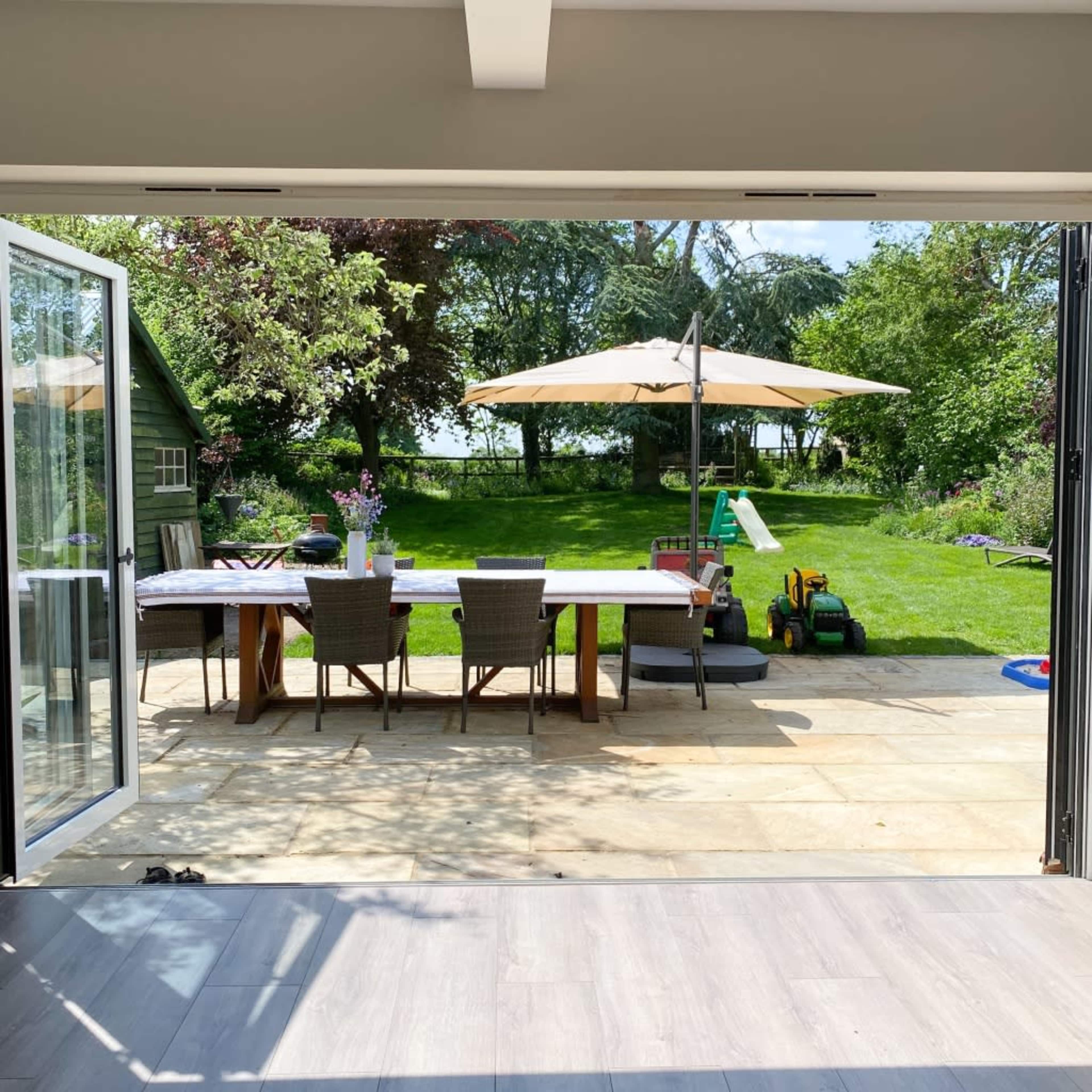 An open patio area with a dining table and chairs, leading to a lush garden featuring a large umbrella, a green lawn, and some children's toys.