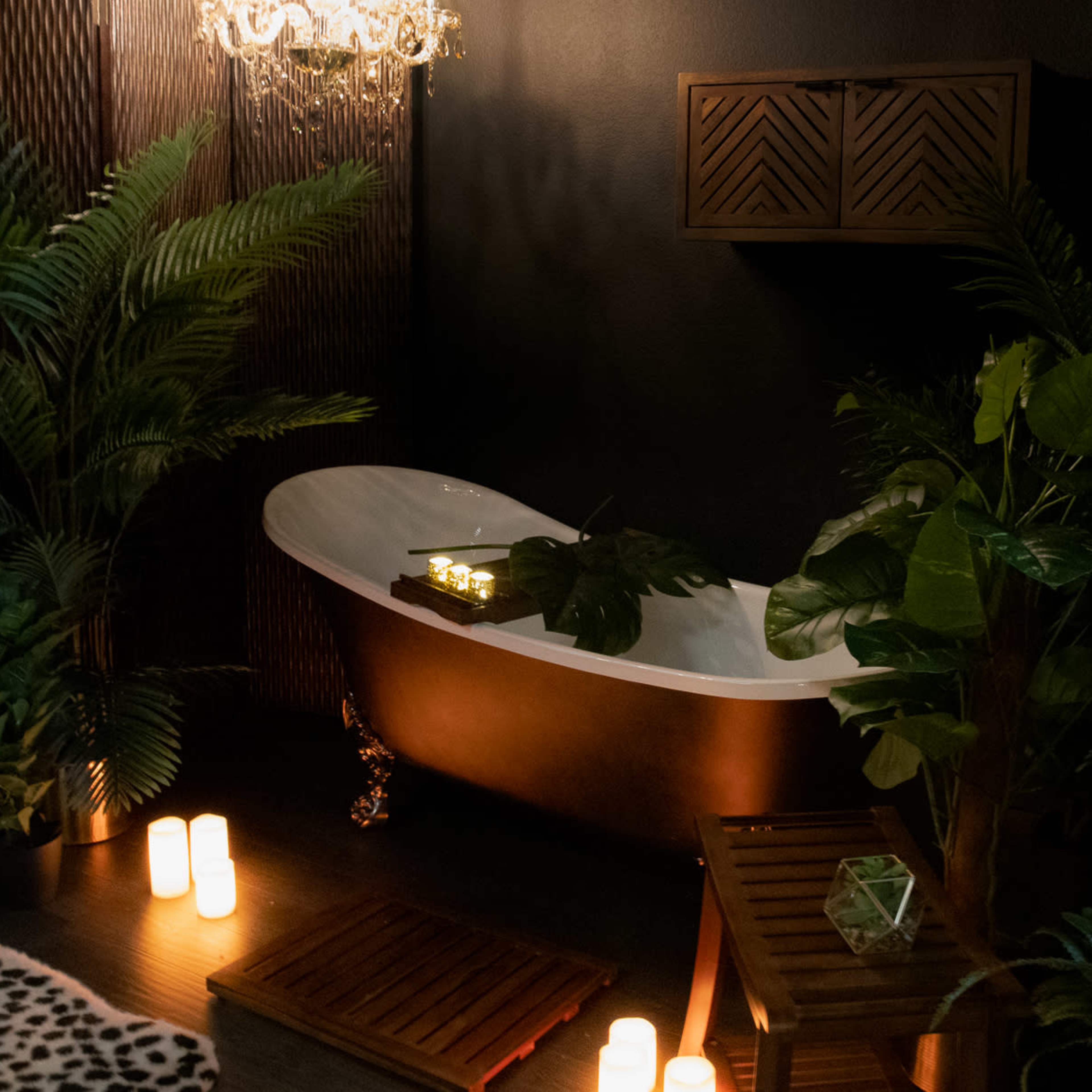 A luxurious bathroom features a freestanding tub surrounded by tropical plants and soft lighting from candles.