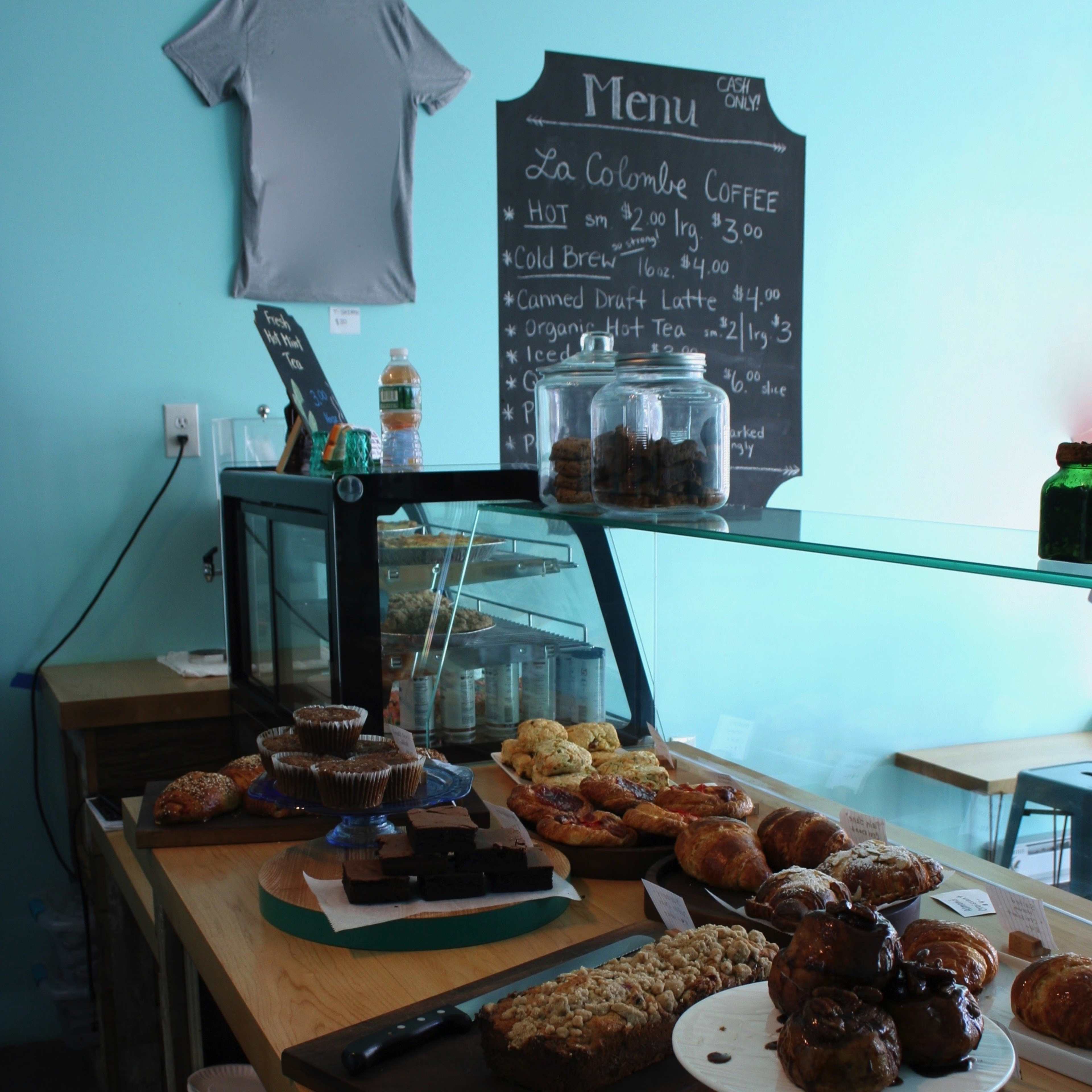 A display of assorted baked goods and coffee options is set on a wooden counter in a café, with a chalkboard menu visible in the background.