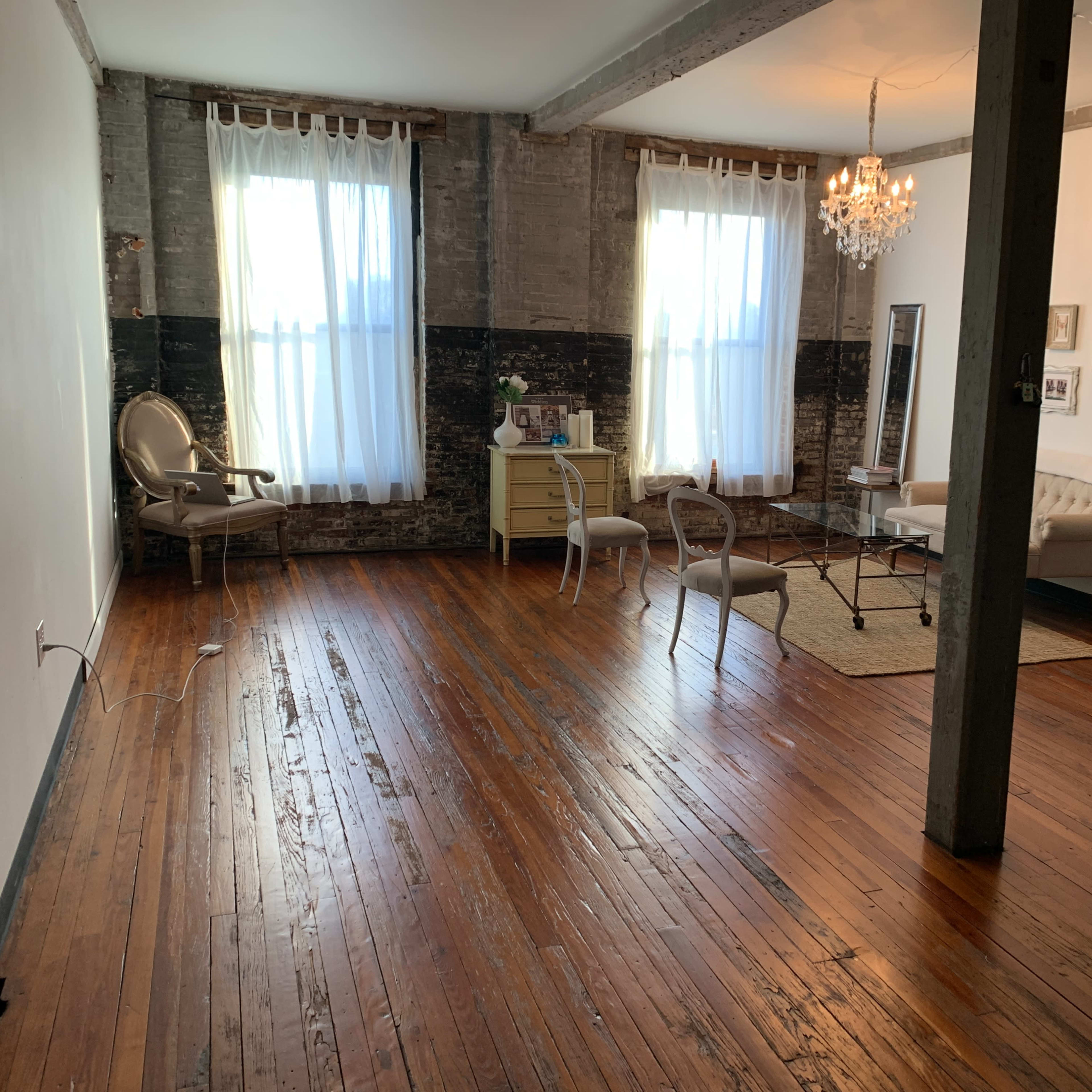 A spacious room with hardwood floors, large windows draped in white curtains, and a mix of vintage furniture including a sofa, chairs, and a chandelier.