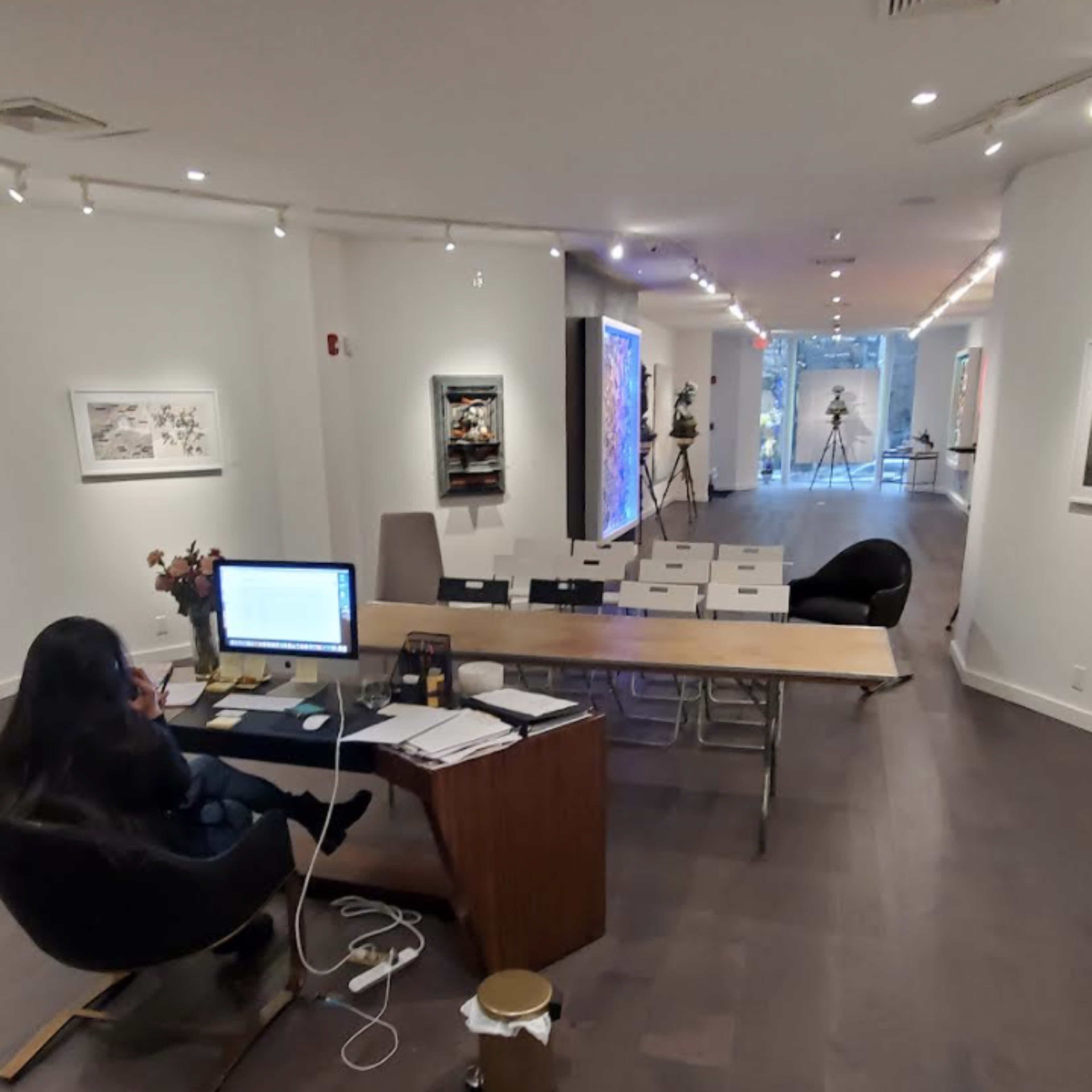 A person works at a desk in an art gallery, with various artworks displayed along the walls and a view of the gallery extending into the background.