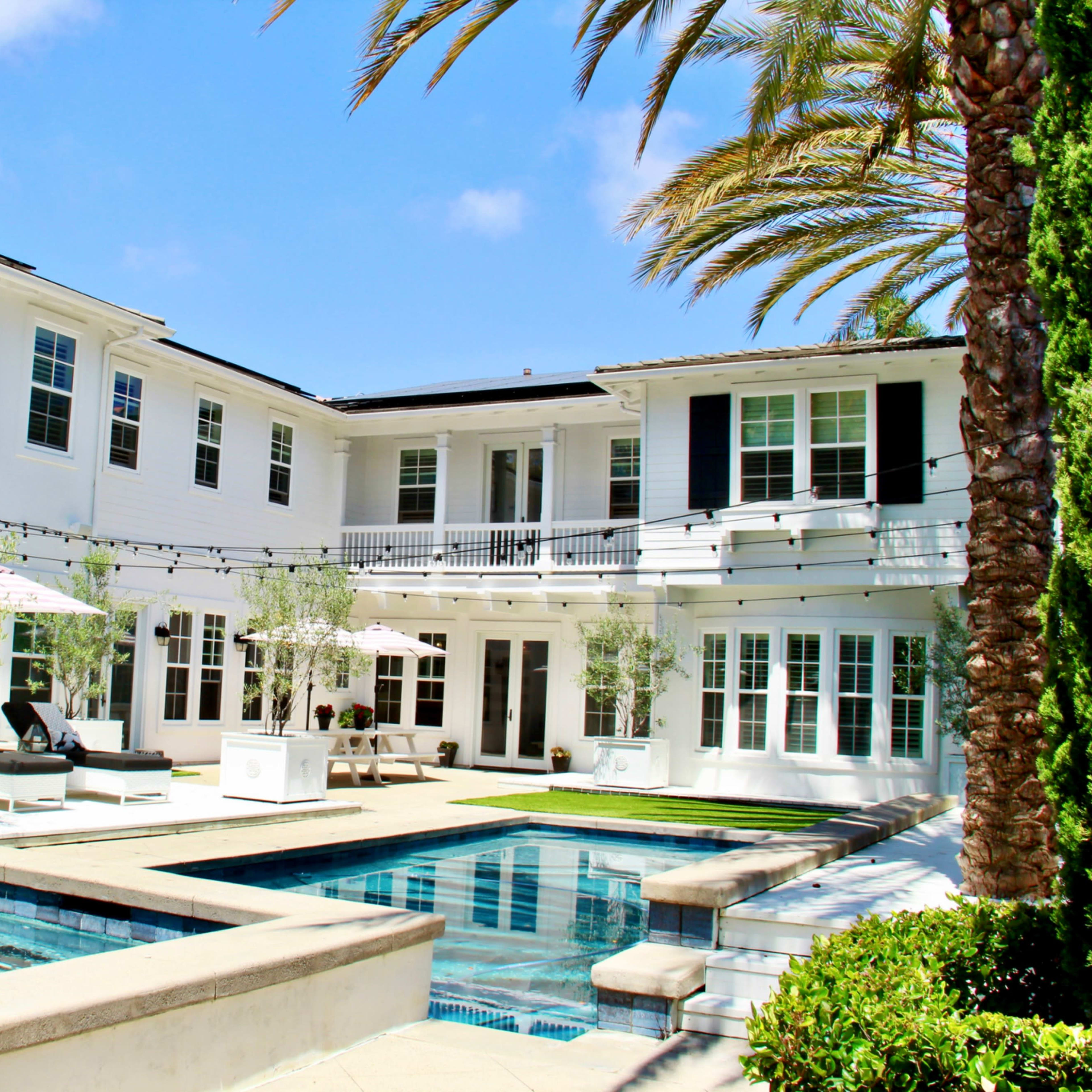 A modern white house with multiple balconies, surrounded by palm trees and featuring two swimming pools in the courtyard.