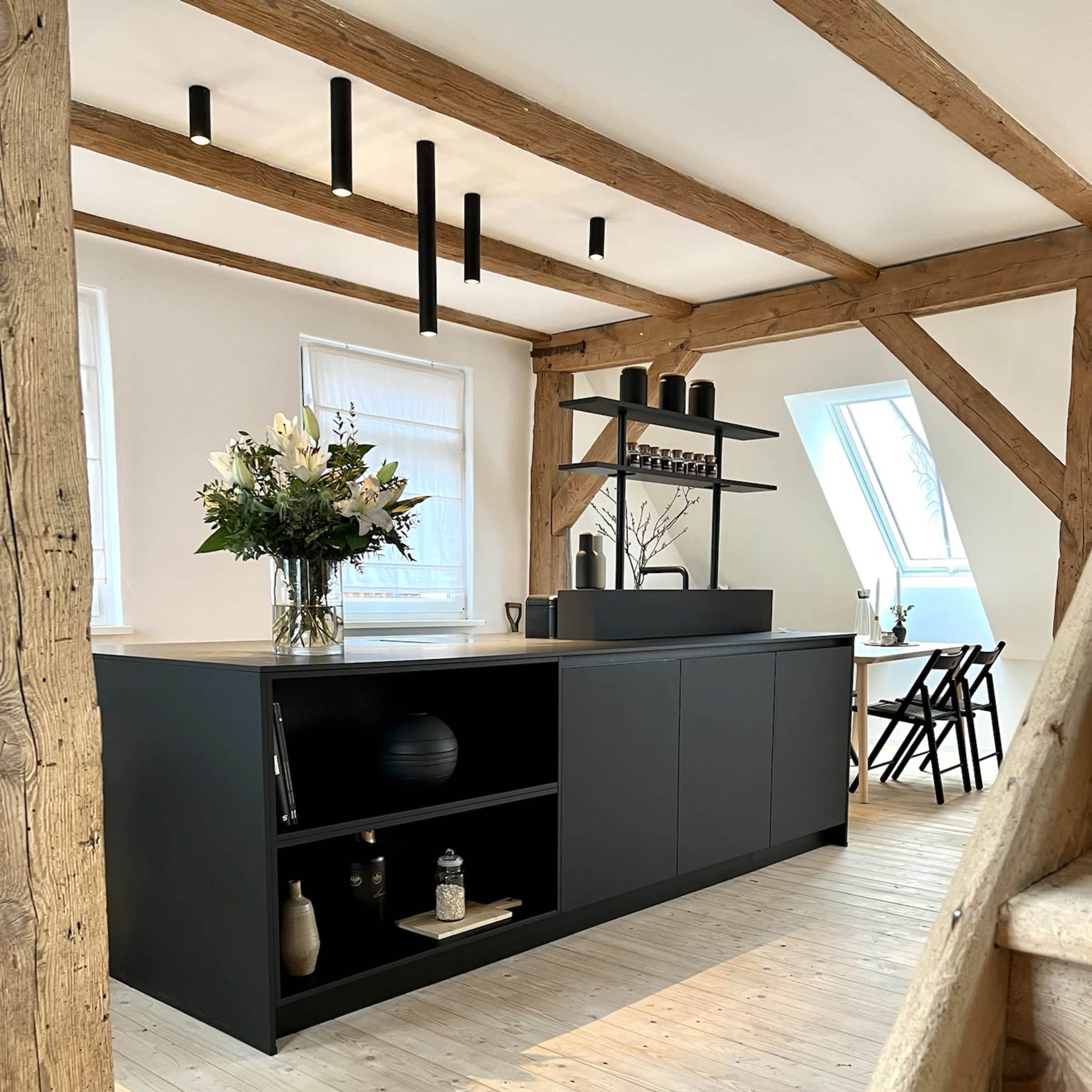 A modern kitchen features a central island with dark cabinetry, wooden beams, and a light wooden floor.