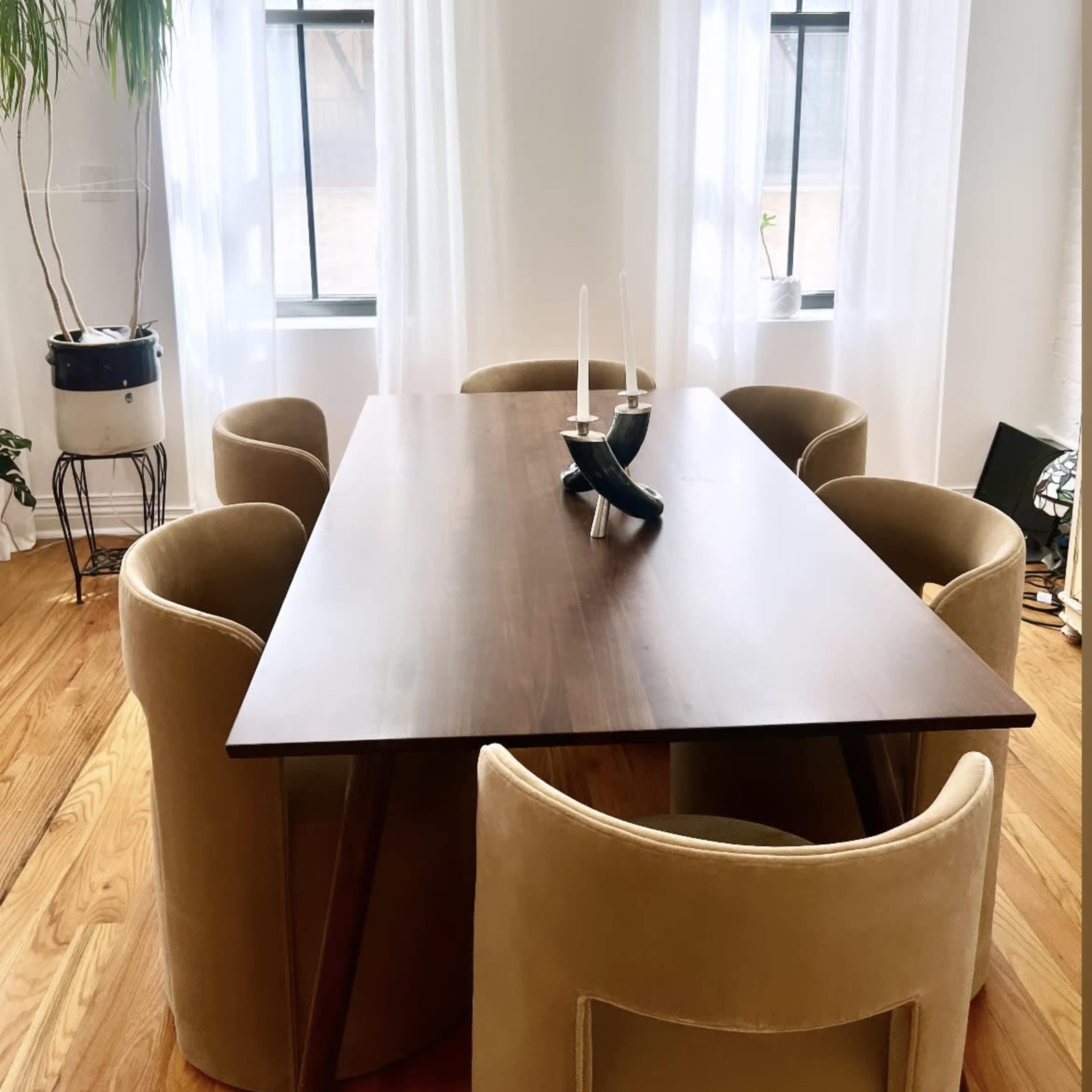 A wooden dining table is surrounded by four upholstered chairs in a brightly lit room with large windows draped in sheer curtains.