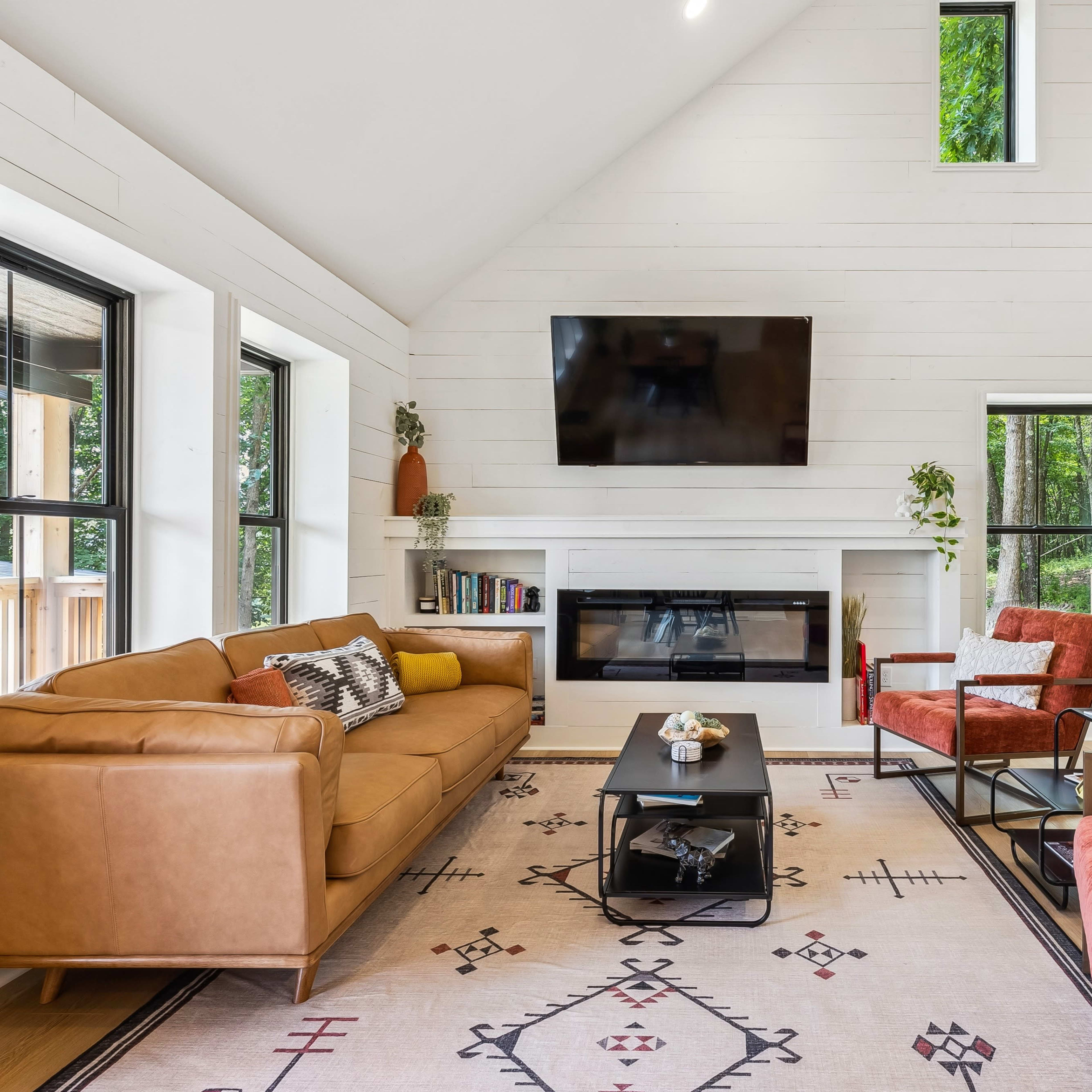 The living room features a brown sofa, two red armchairs, a black coffee table, and a mounted television above a modern fireplace, with large windows allowing natural light to enter.