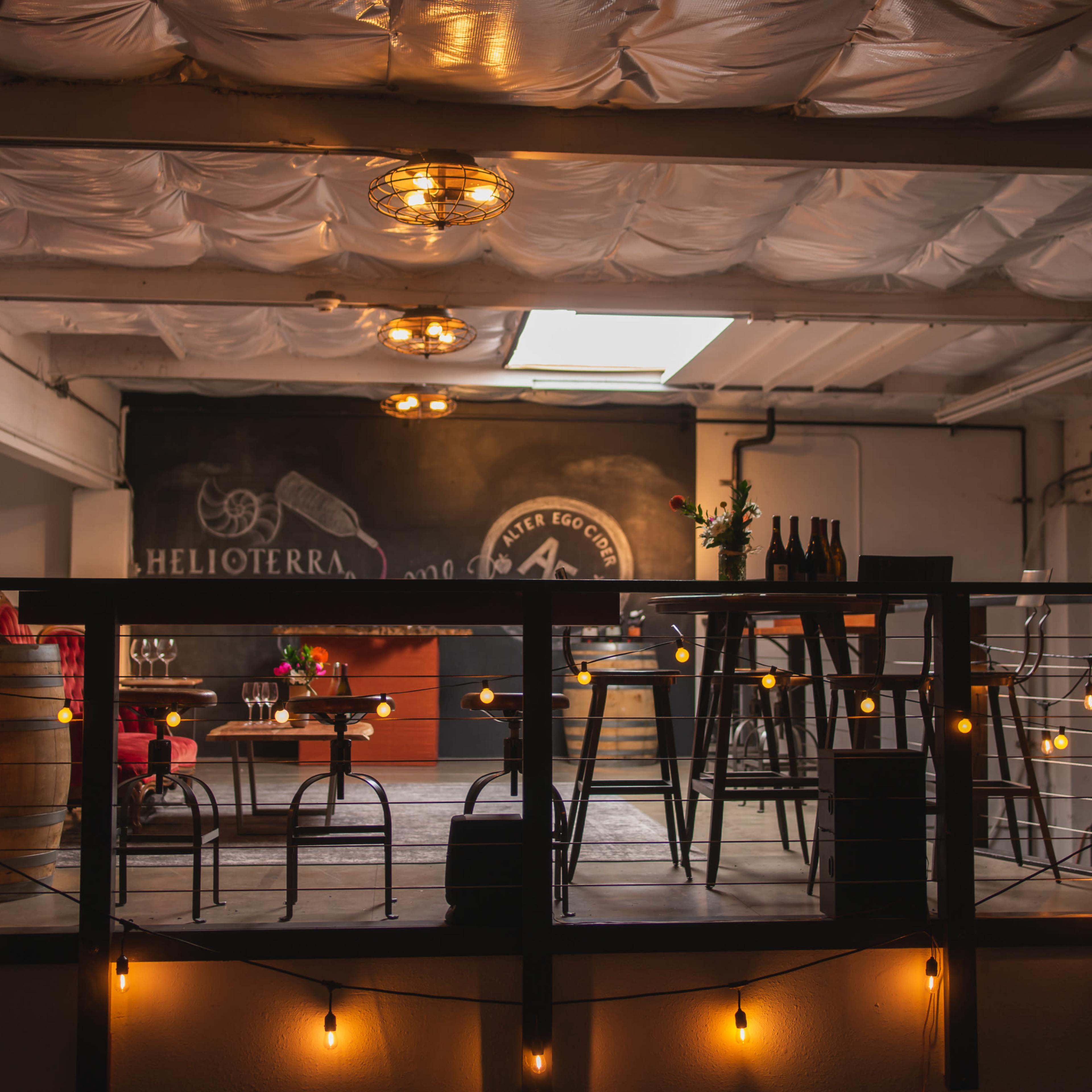A cozy upper level of a bar or wine tasting room featuring wooden furniture, string lights, and a chalkboard background.