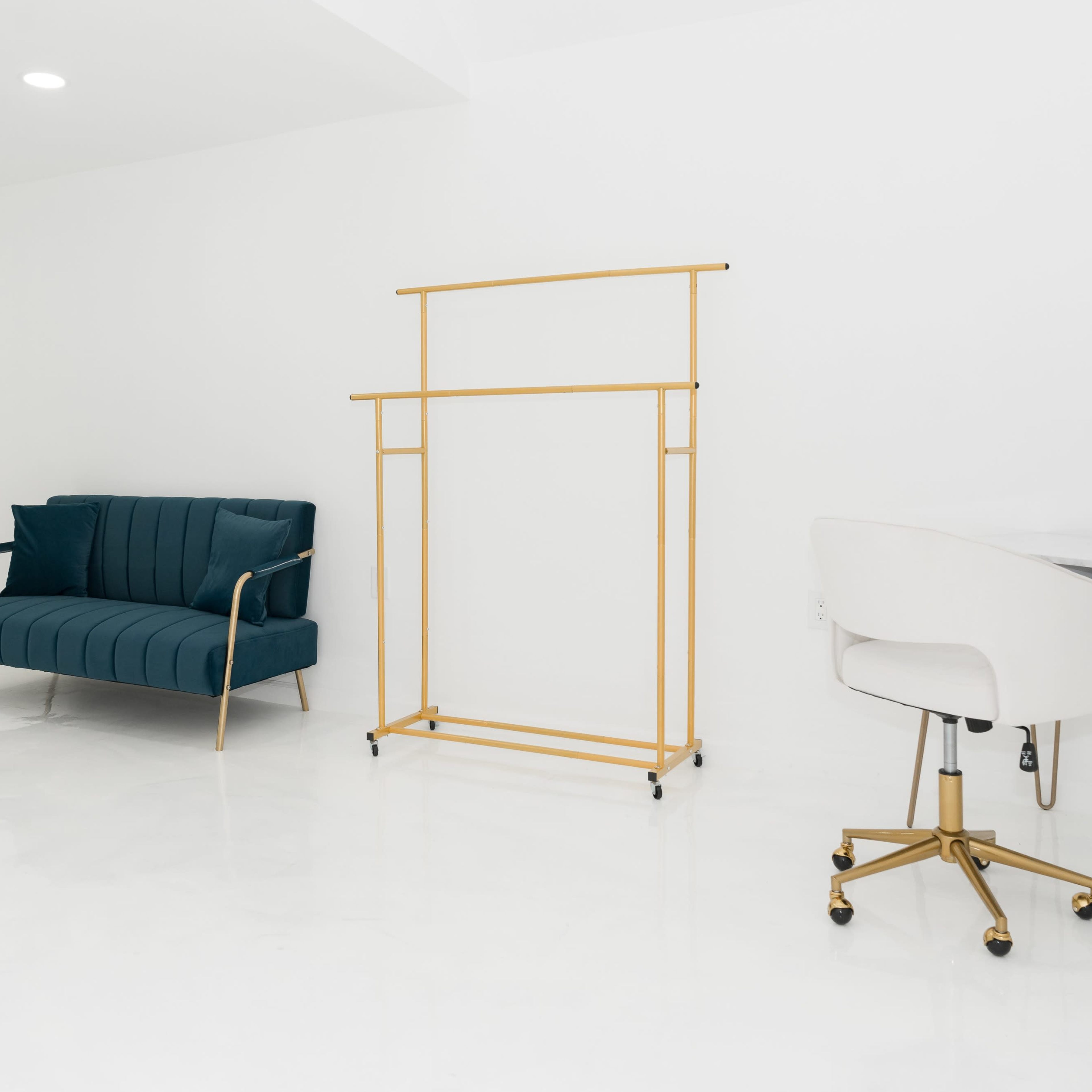 A minimalist room with a teal sofa, a gold clothing rack, a white desk chair, and a water cooler.