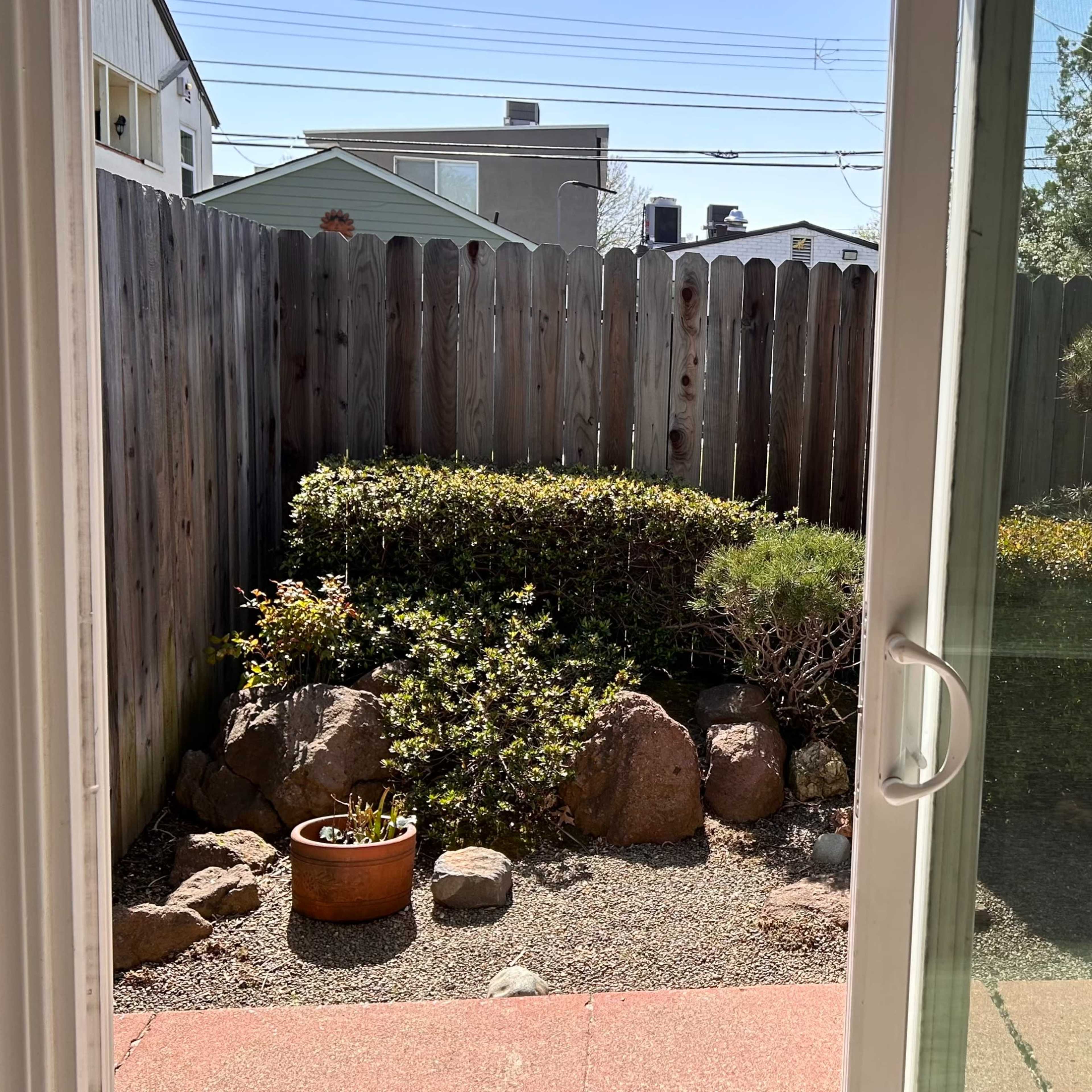 A view through a glass door into a small backyard garden with a landscaped area featuring rocks, shrubs, and a couple of chairs.