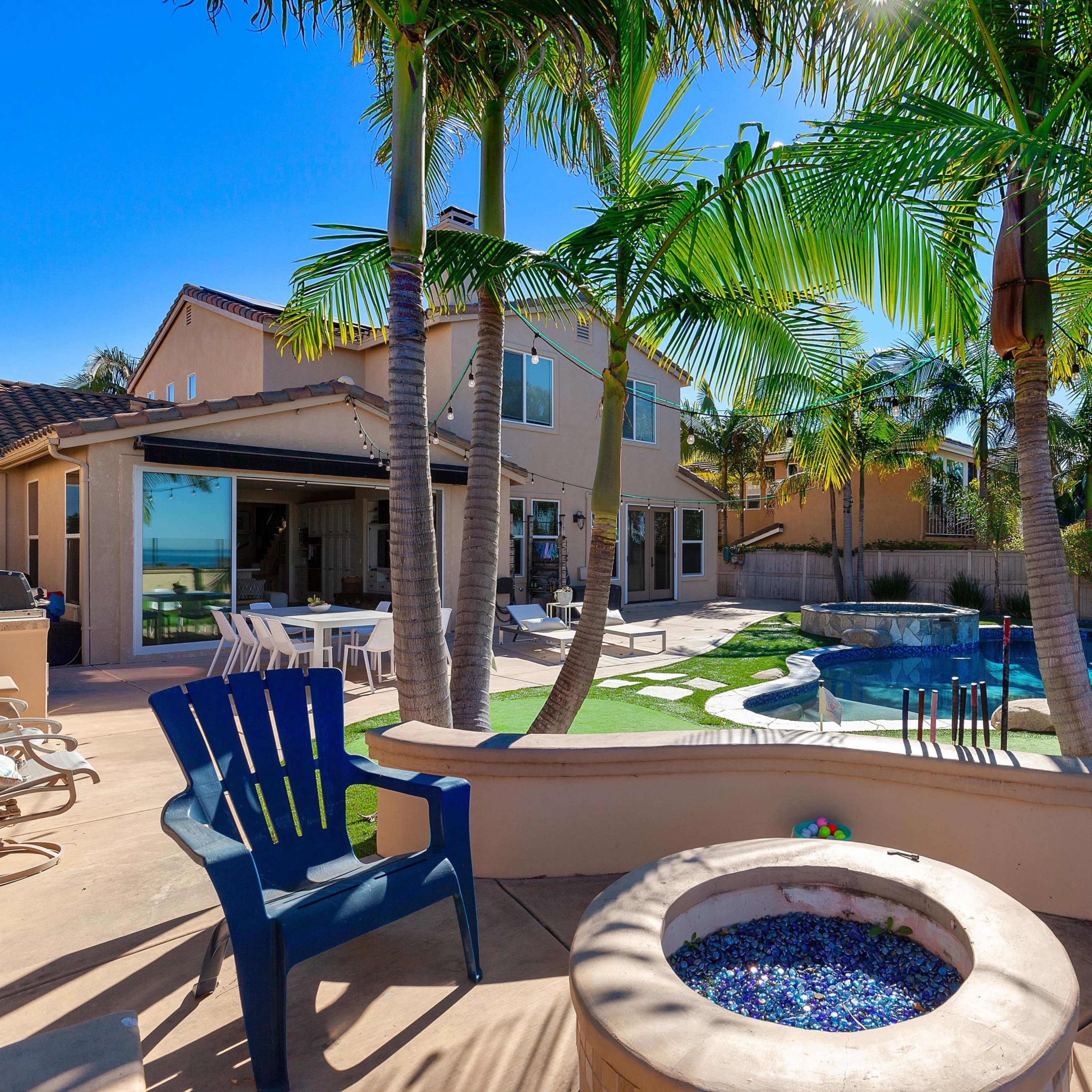 A backyard with a swimming pool, palm trees, and a fire pit, surrounded by a house and patio furniture.