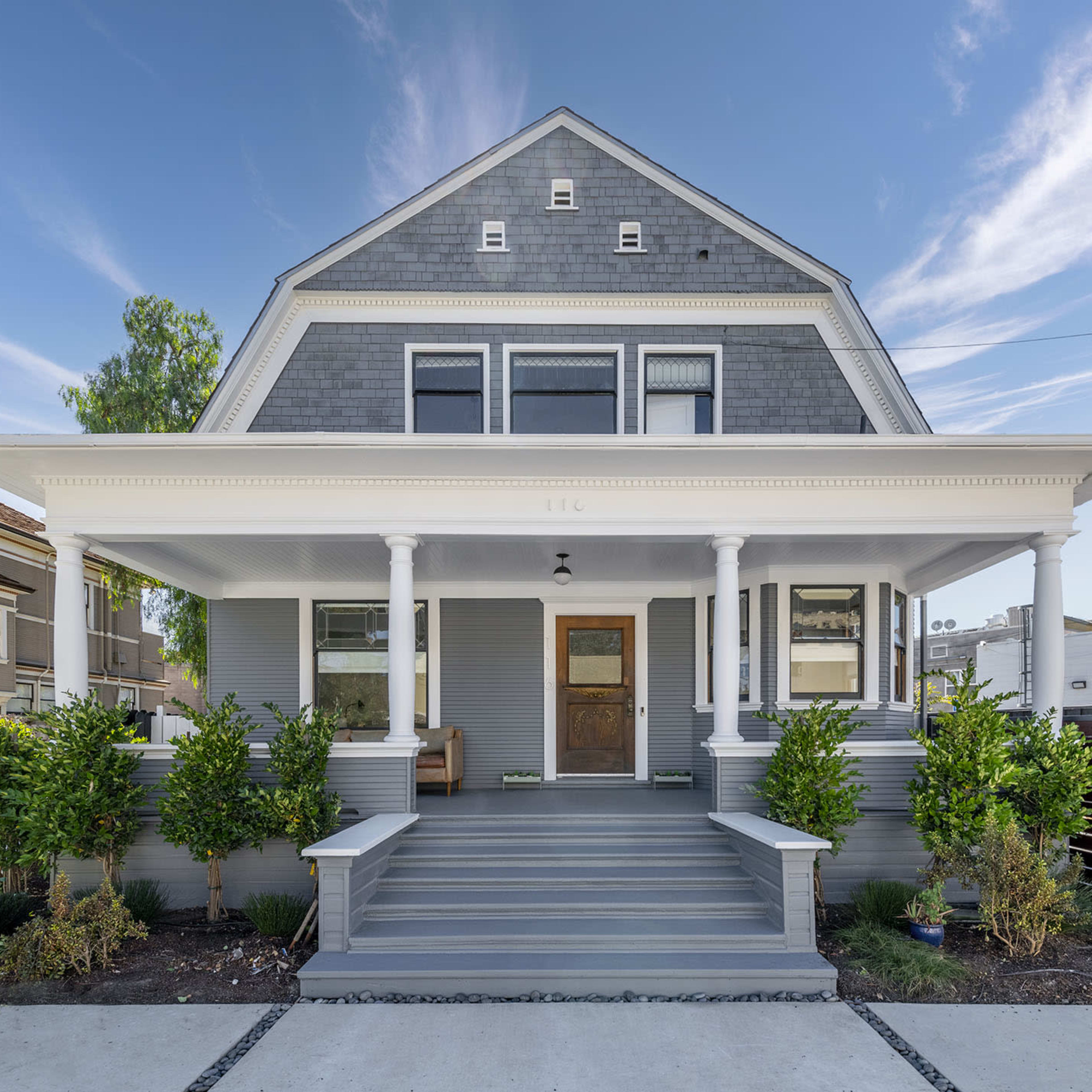A gray-painted house with a wide porch and columns is surrounded by neatly trimmed bushes and landscaping.