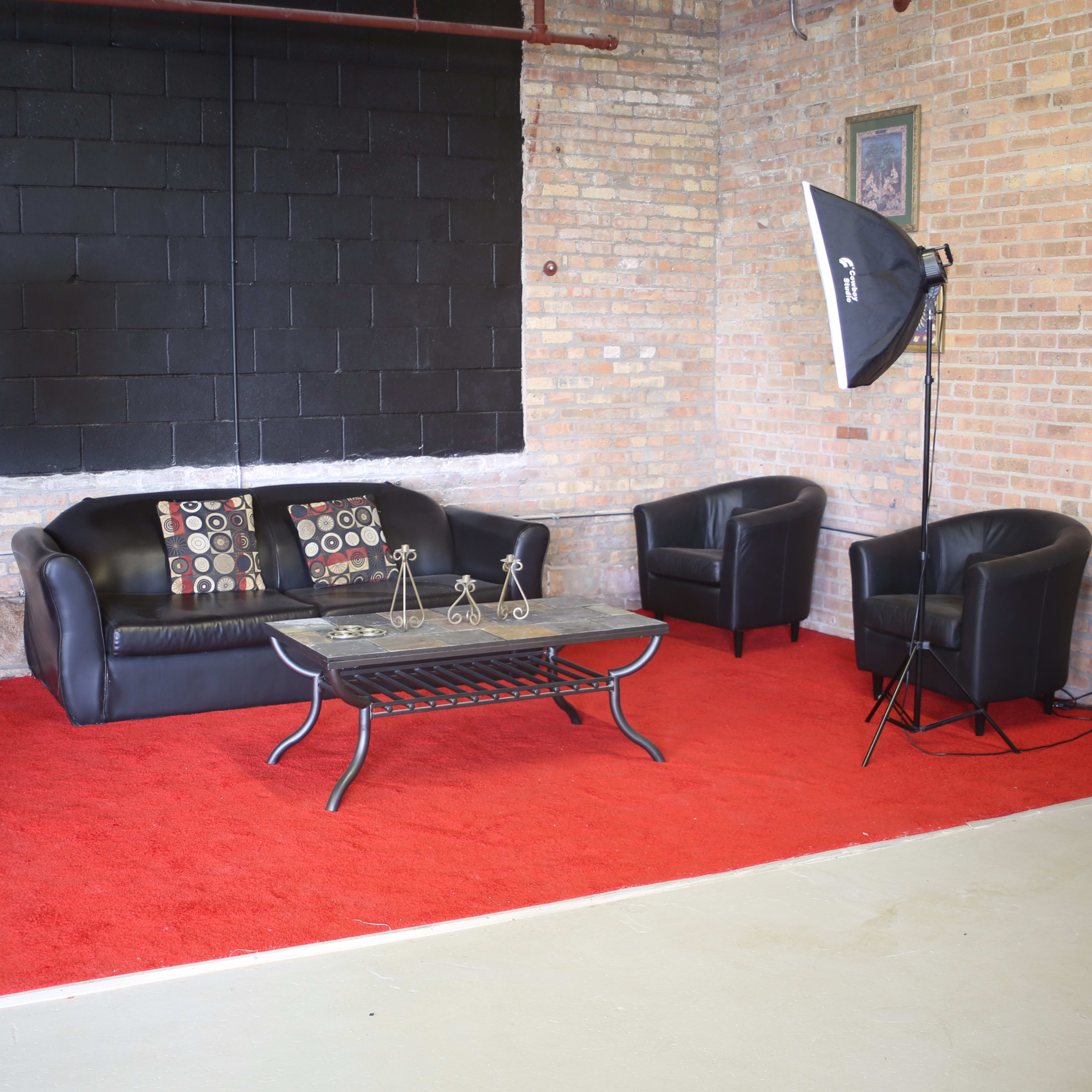 A seating area with a black sofa, two matching armchairs, a coffee table, and professional lighting equipment on a red carpeted floor against a brick wall.
