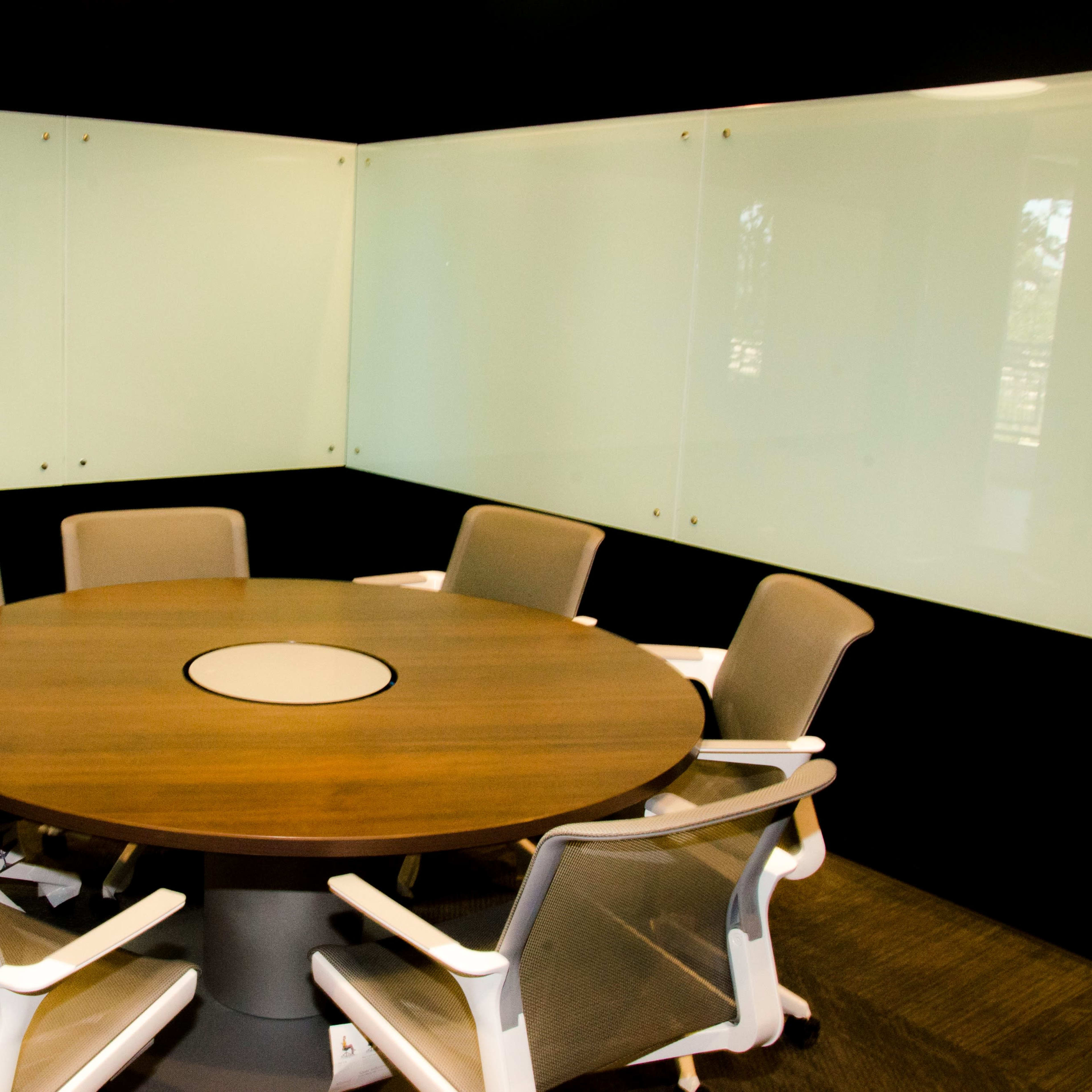 A circular wooden conference table surrounded by six ergonomic chairs in a conference room with glass walls.