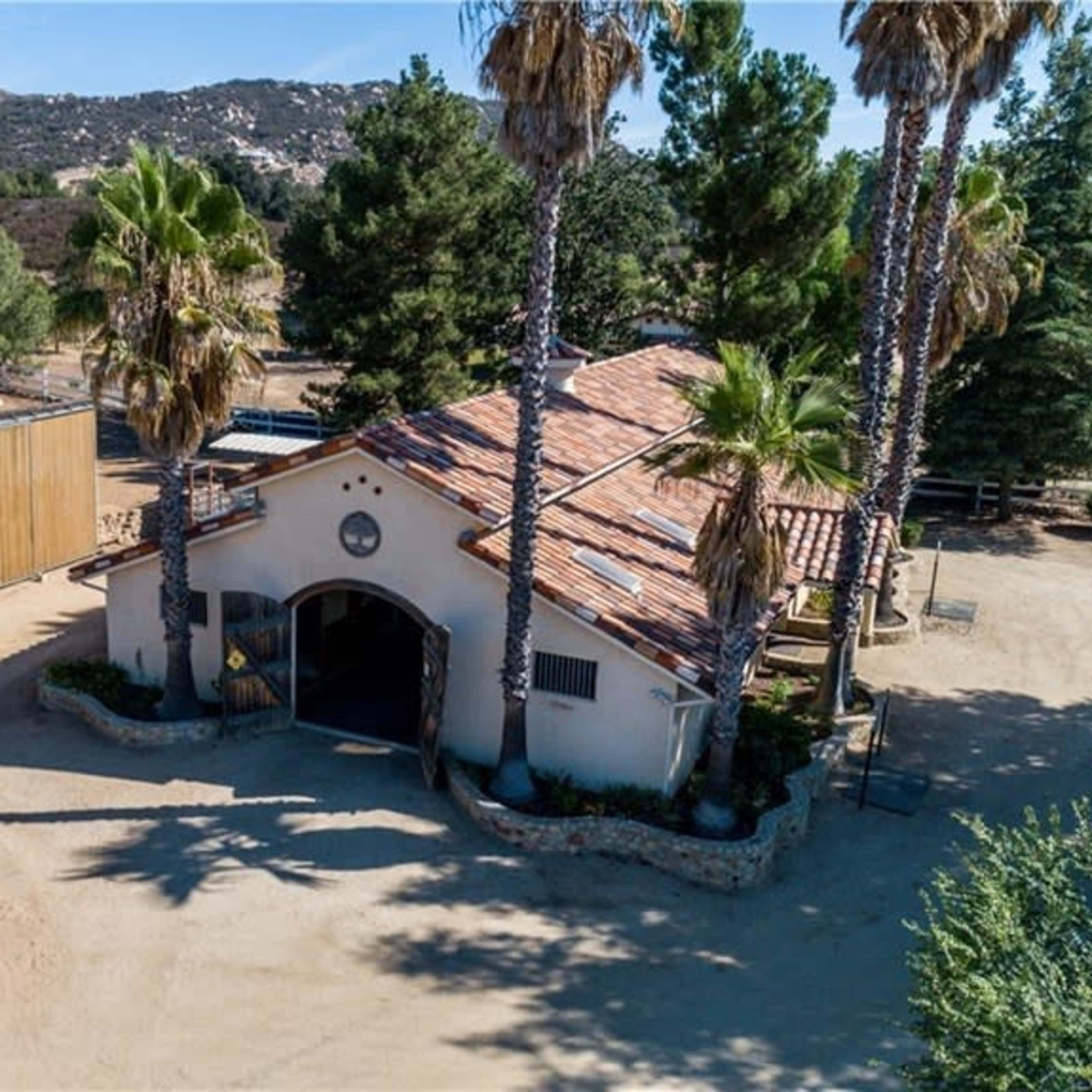 A single-story building with a red-tiled roof surrounded by palm trees and dirt paths, set against a mountainous landscape.