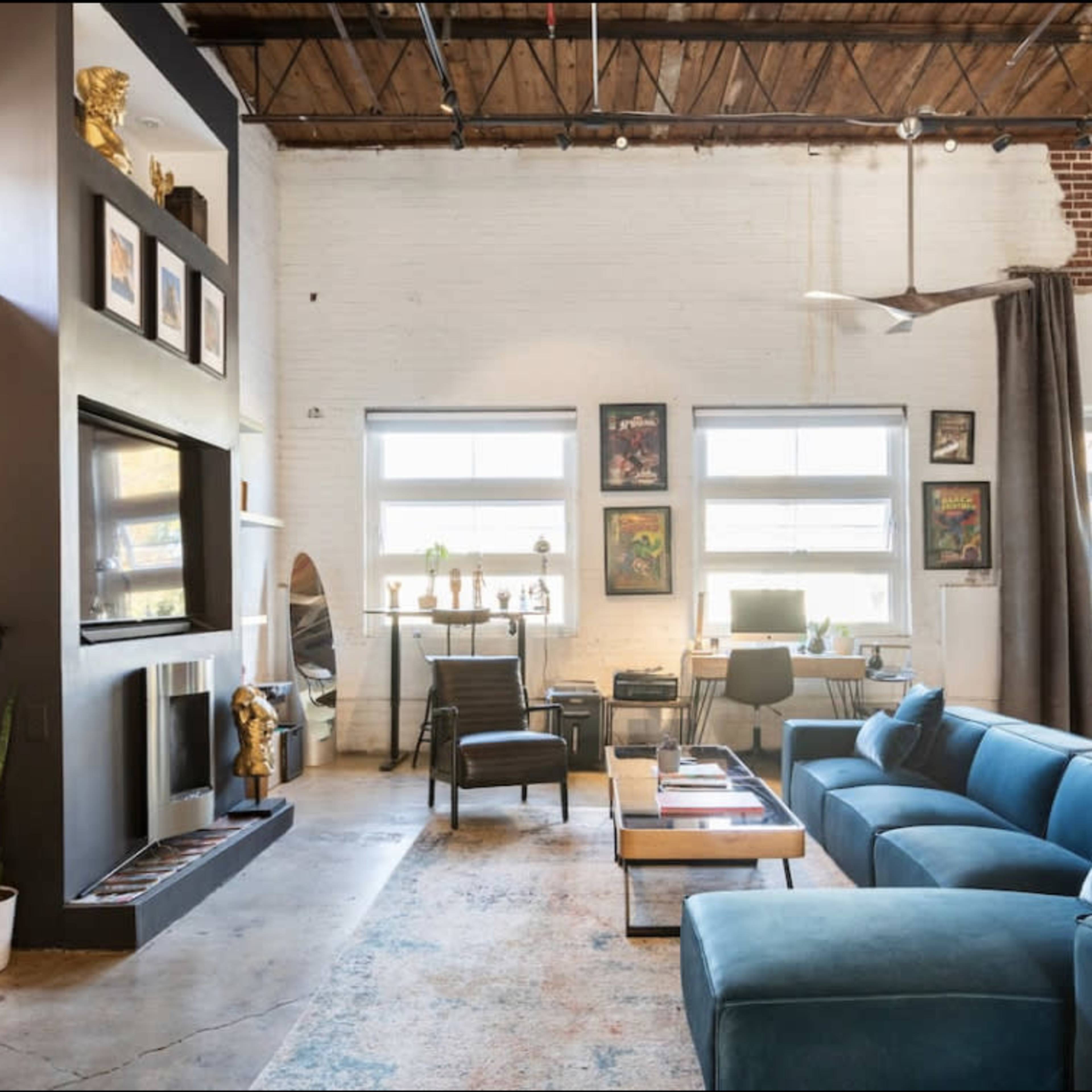 A spacious living area with a blue sectional sofa, large windows, and a mix of modern and vintage decor, including artwork and plants.