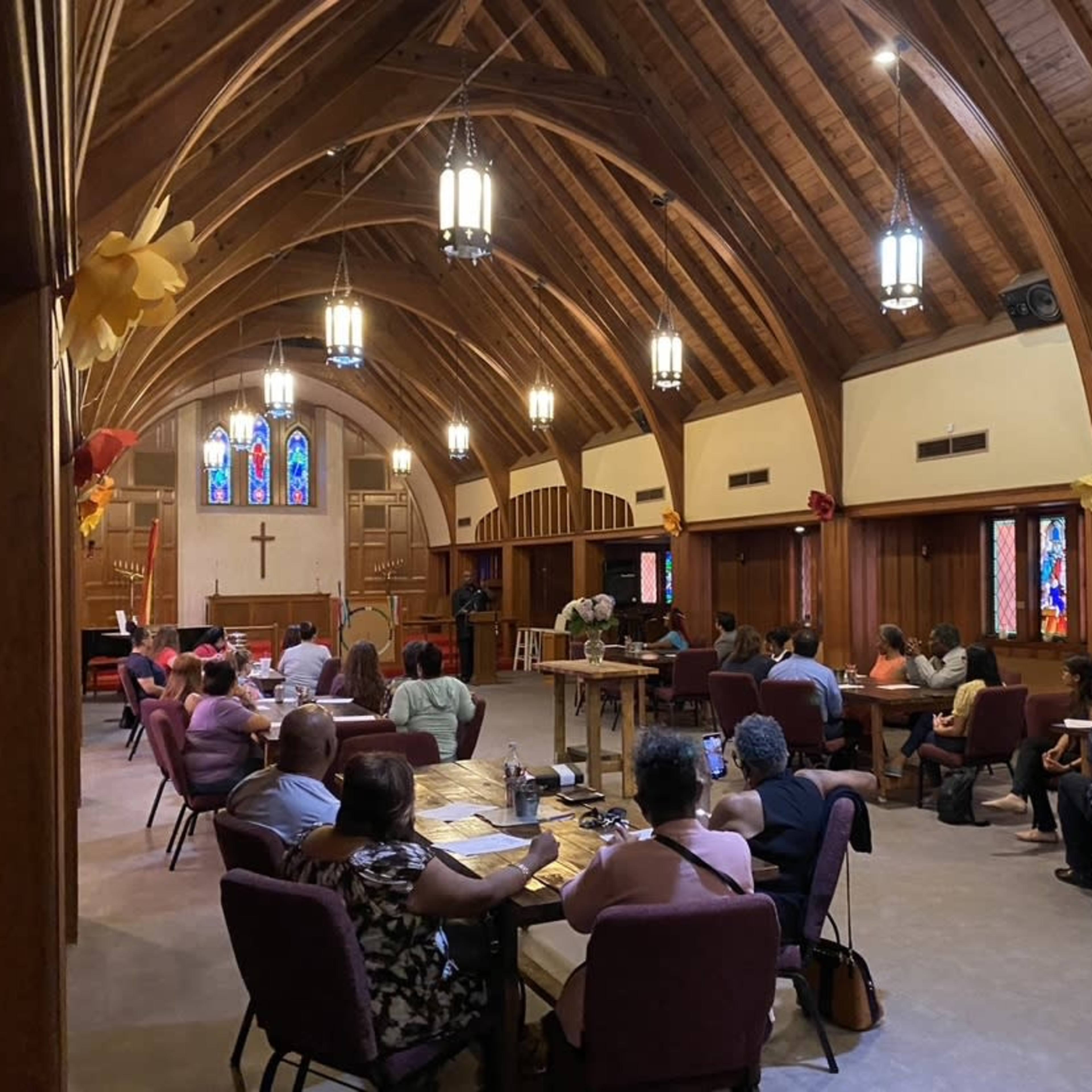 A group of people is seated at tables in a church with wooden beams, stained glass windows, and decorative elements.