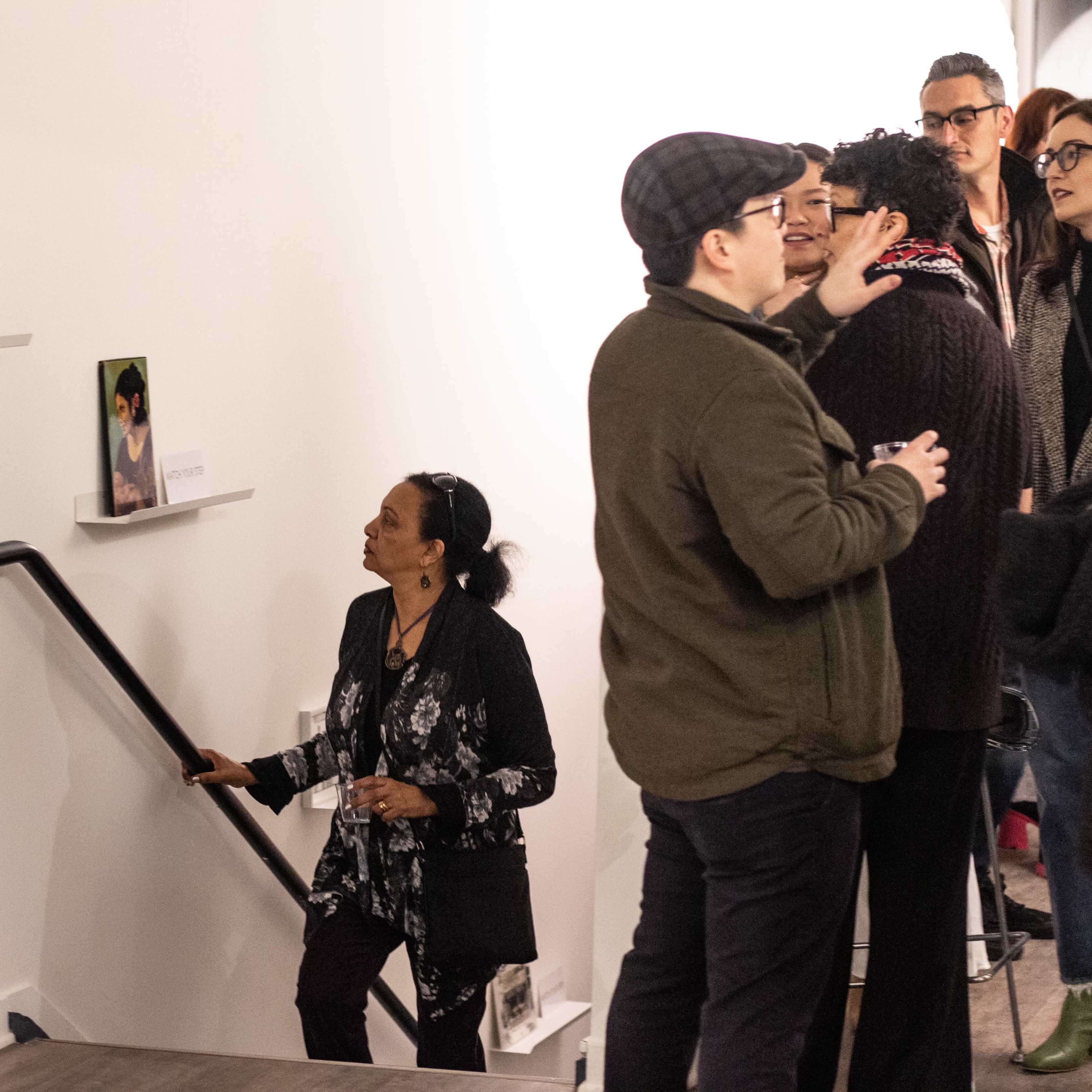 A woman ascends a staircase while a group of people engages in conversation nearby, with various artworks displayed on the walls.