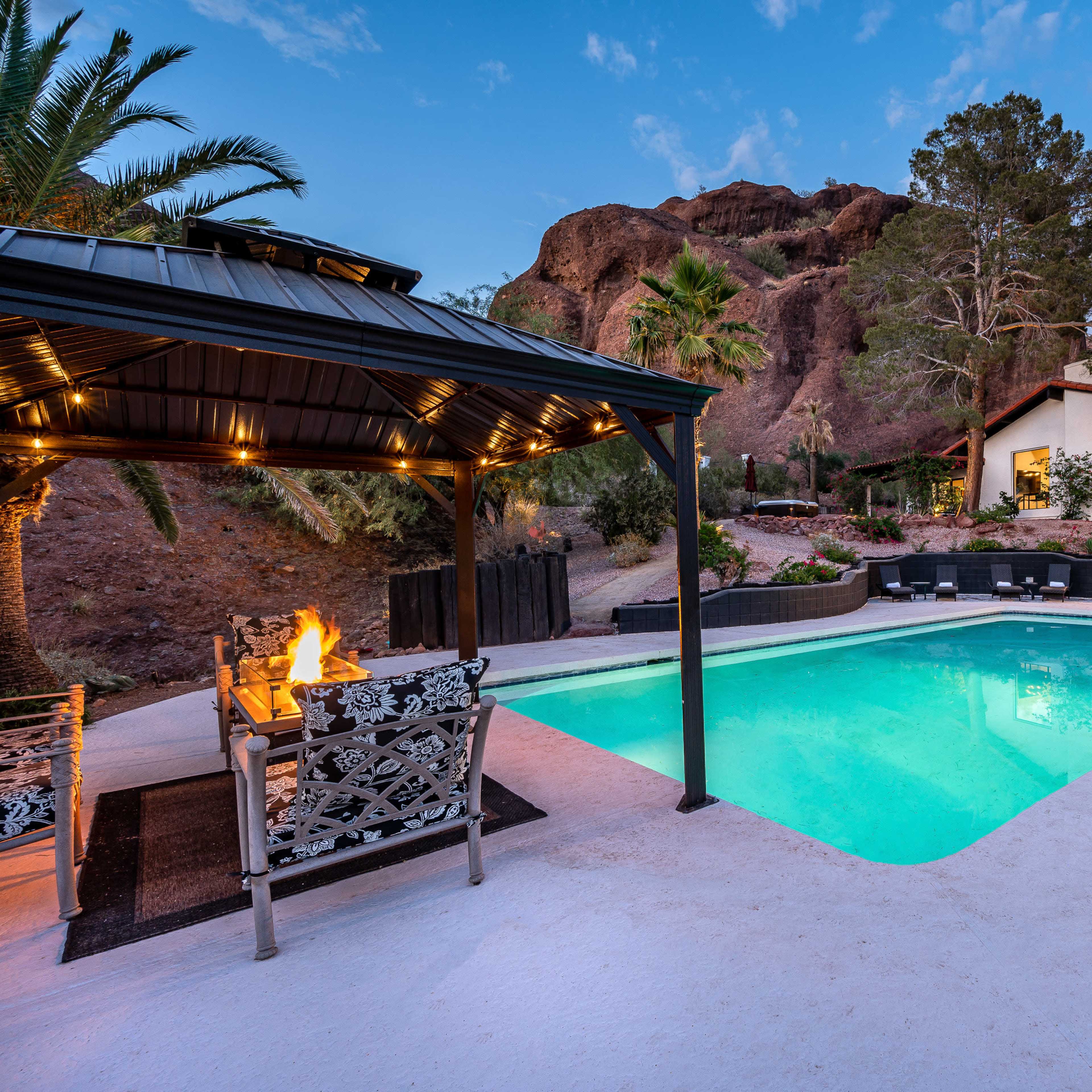 A swimming pool with a gazebo and seating area surrounded by palm trees and rocky terrain.