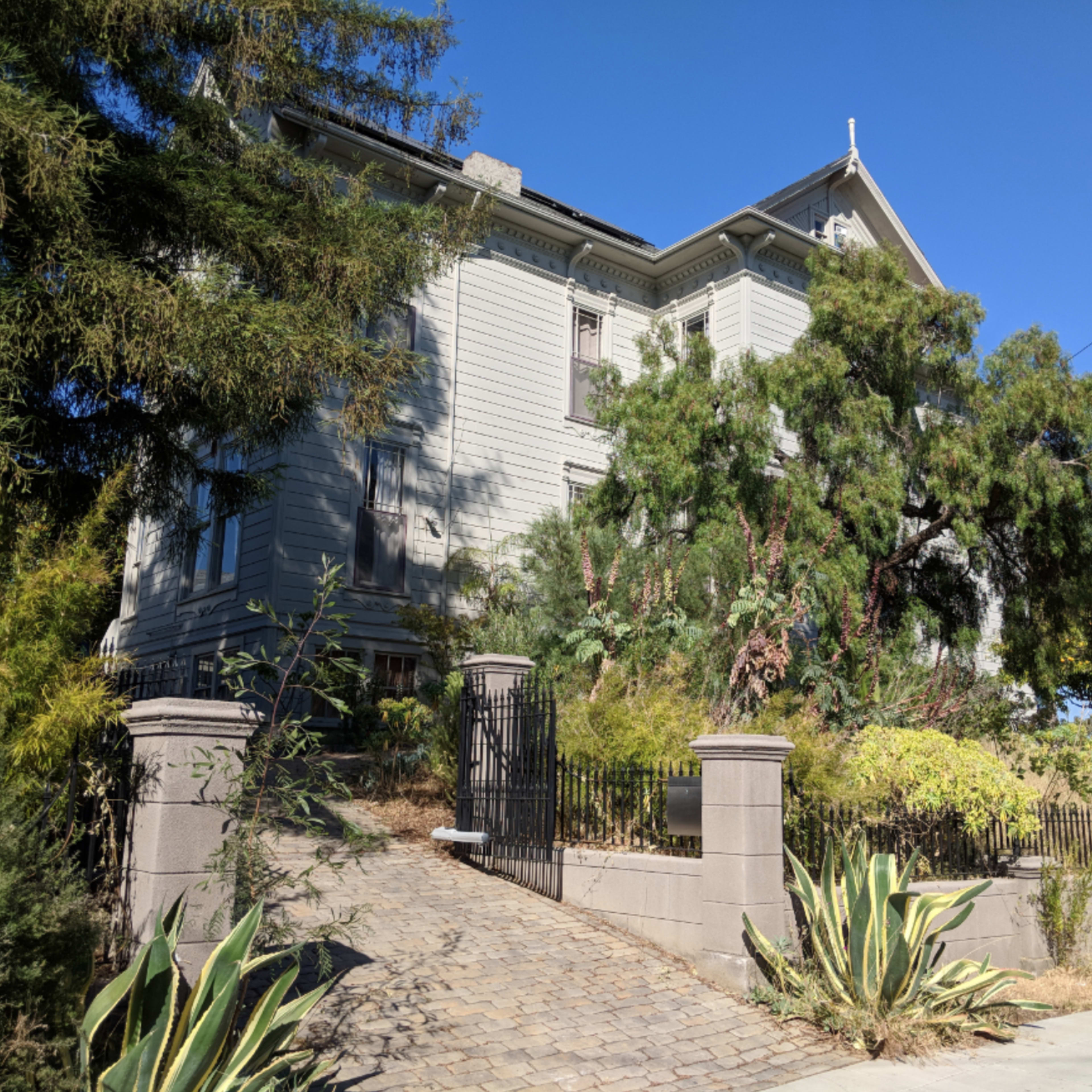 A large, light-colored Victorian house is surrounded by greenery and features a gated entrance with a cobblestone pathway leading to it.