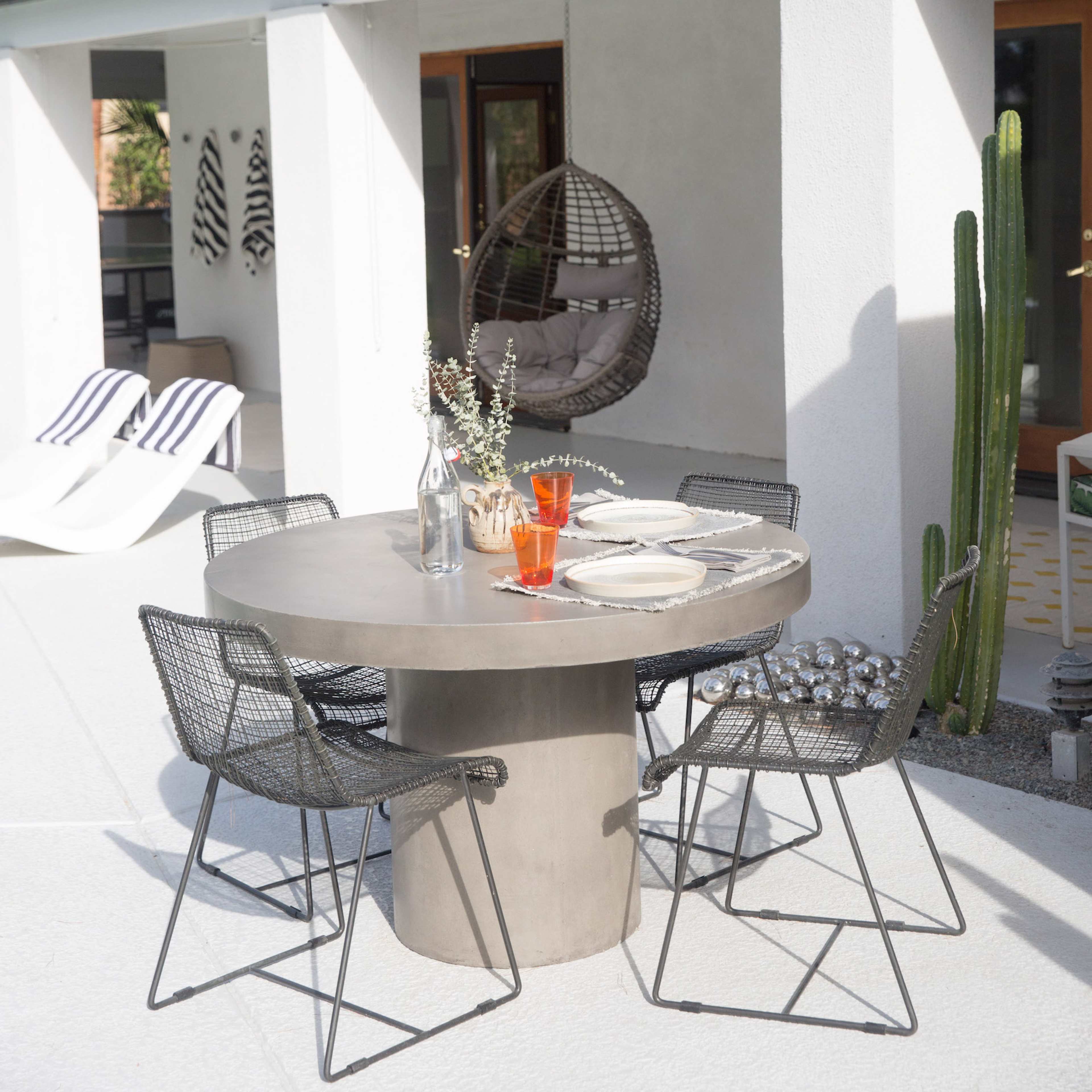 A circular concrete dining table surrounded by black wire chairs is set for a meal on a patio with palm trees in the background.