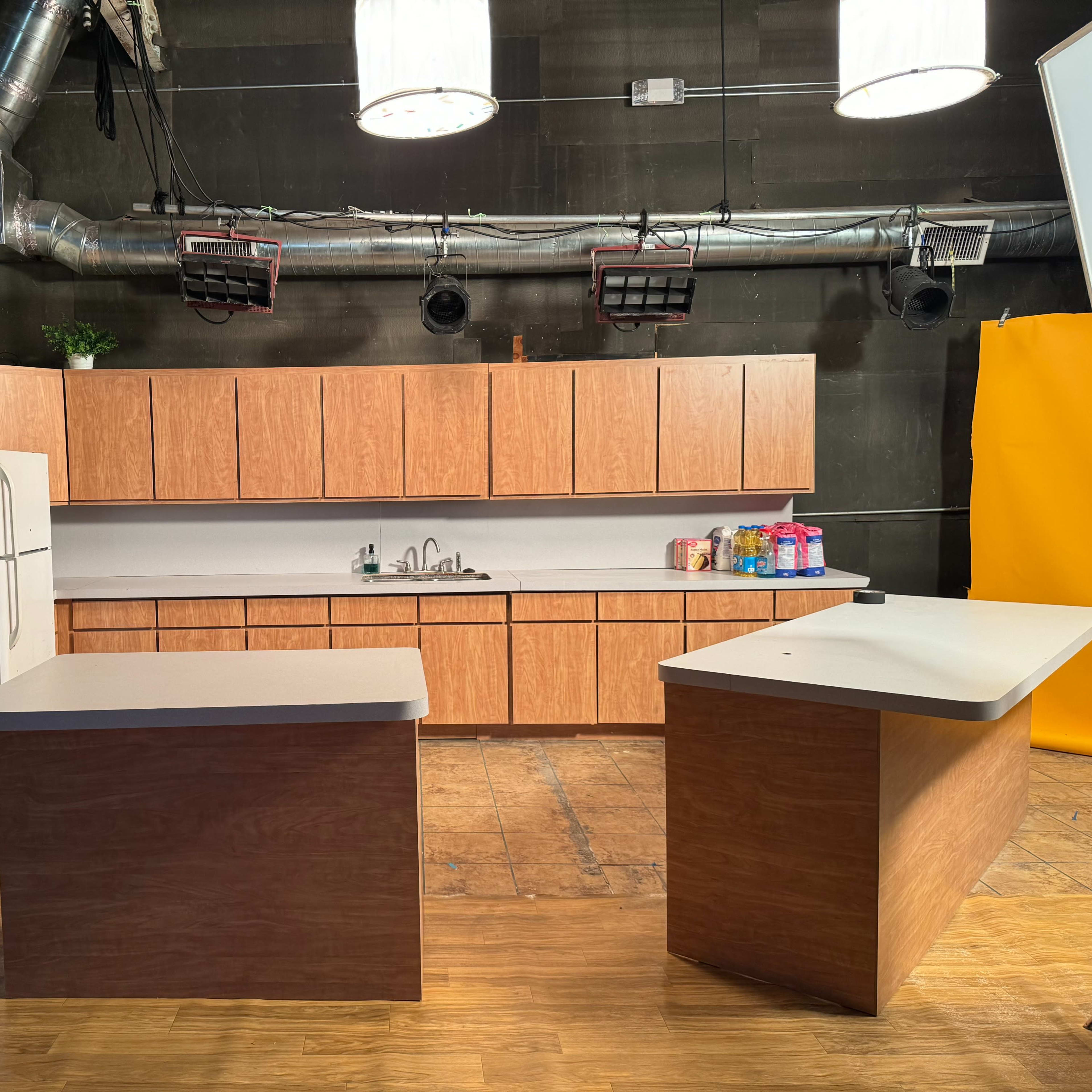 A kitchen set features wooden cabinetry, a refrigerator, and a central island under bright studio lights.