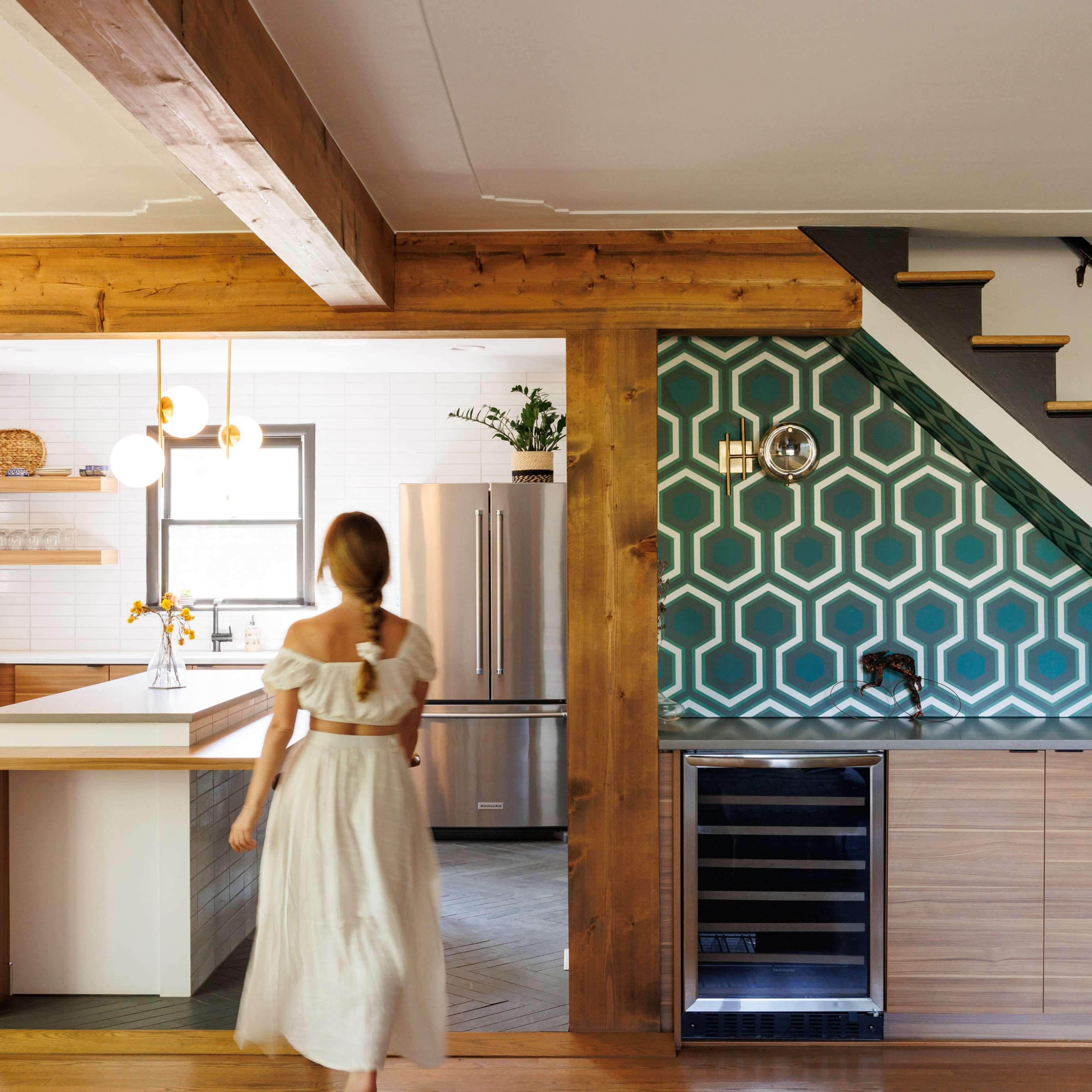 A woman in a white dress walks through a kitchen with a hexagonal tile backsplash and wooden beams along the ceiling.