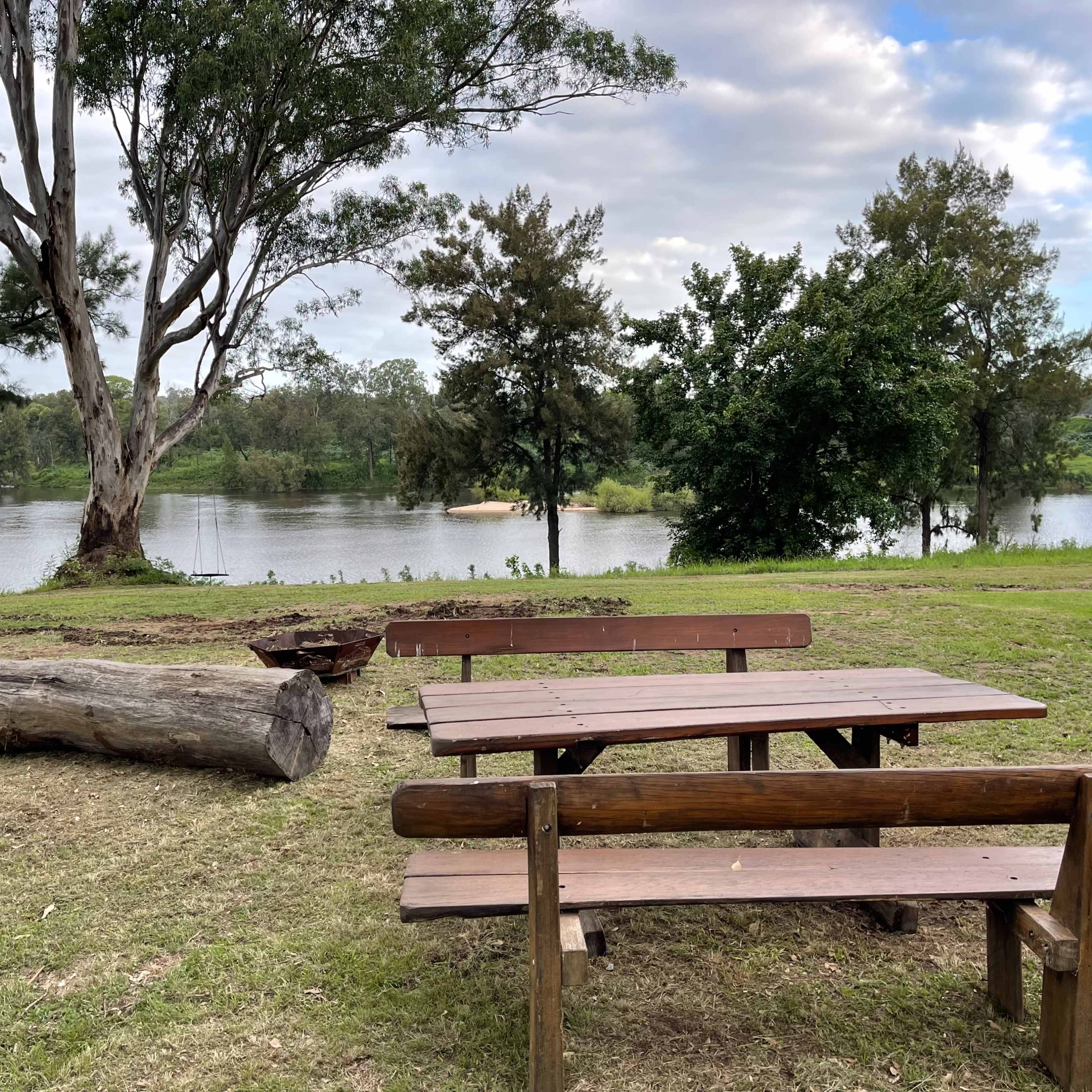 A picnic area with wooden tables and a large log overlooks a calm river surrounded by trees.