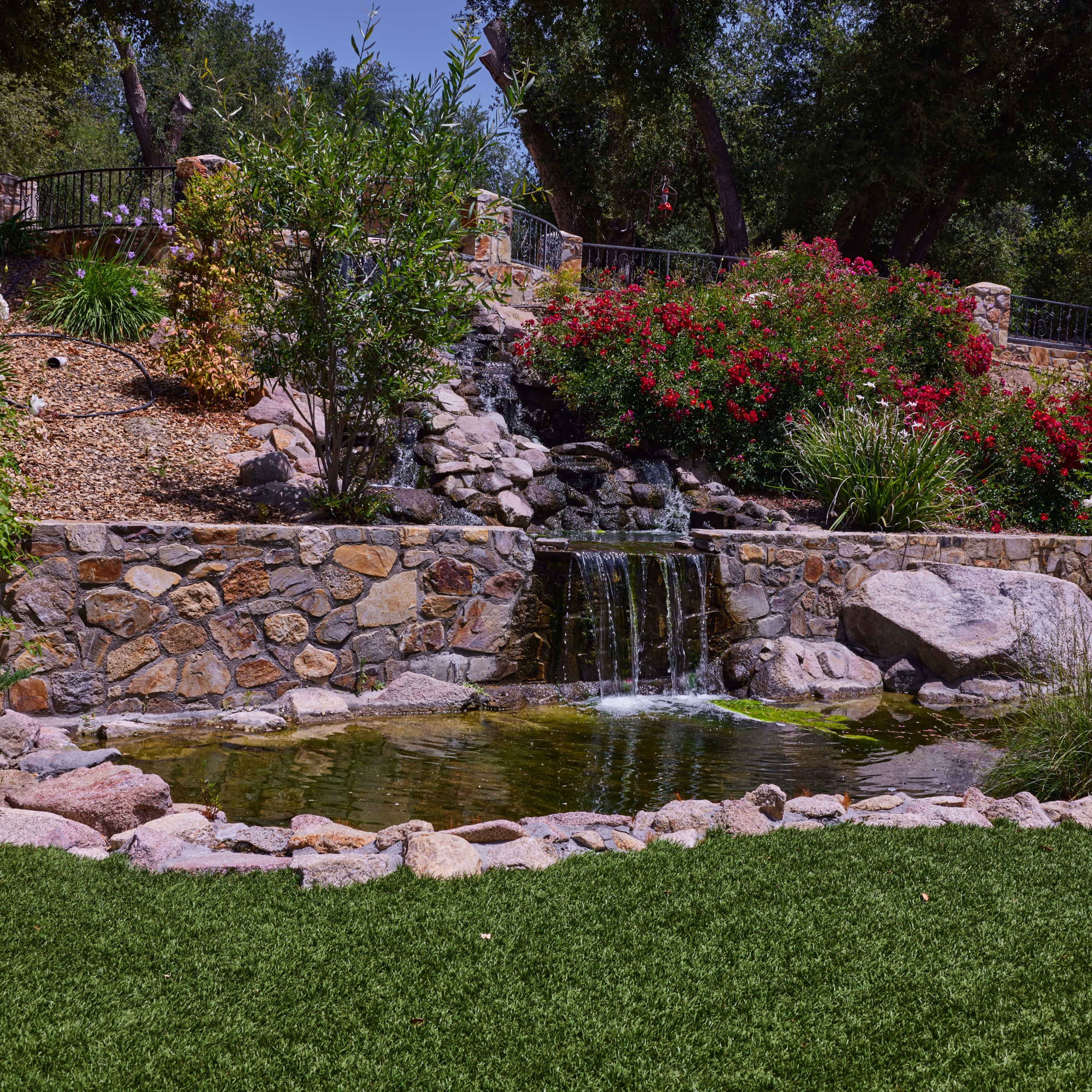 A stone pond with a small waterfall, surrounded by lush greenery and colorful flowers set against a hillside.