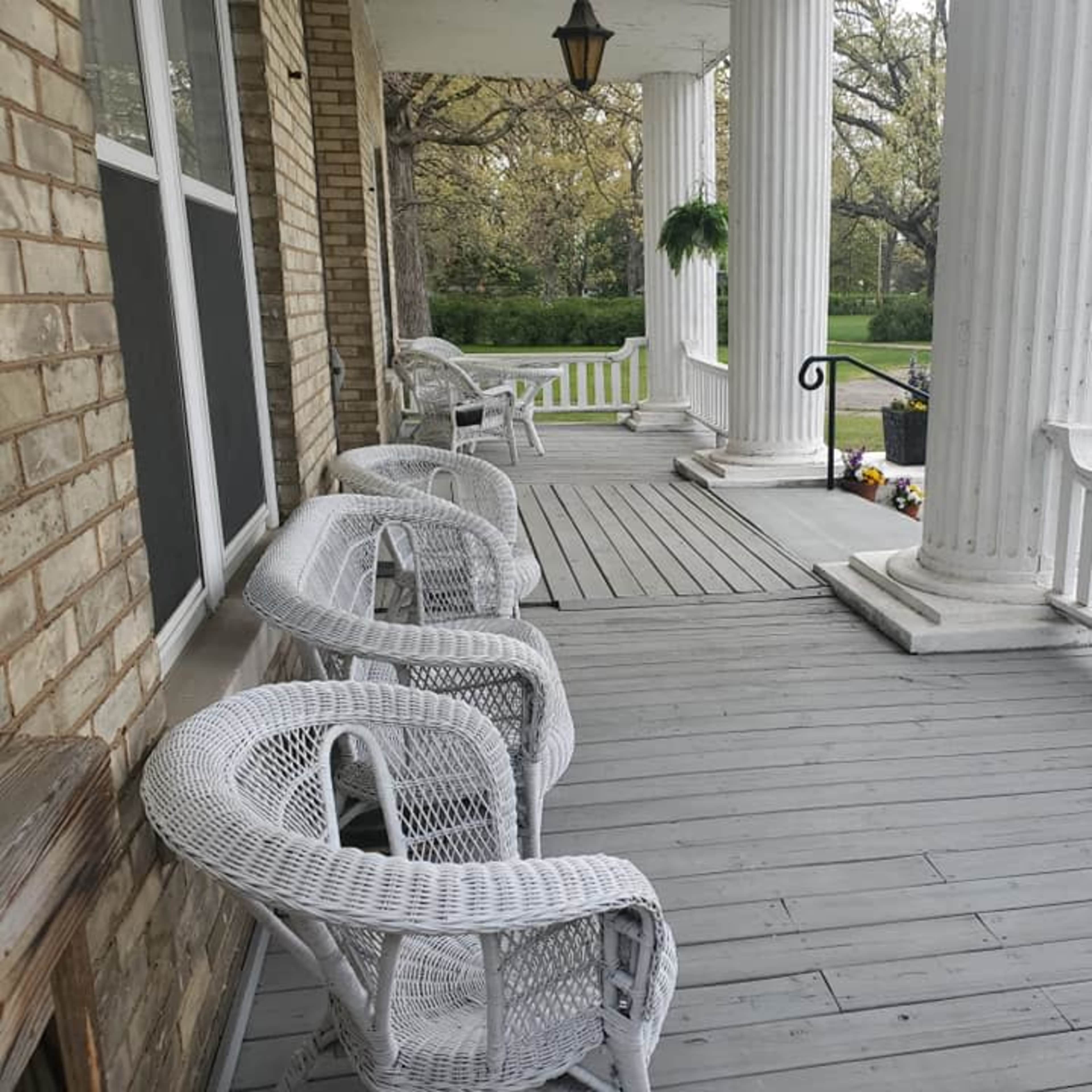 A porch features white wicker chairs arranged near tall columns and a view of a green lawn.