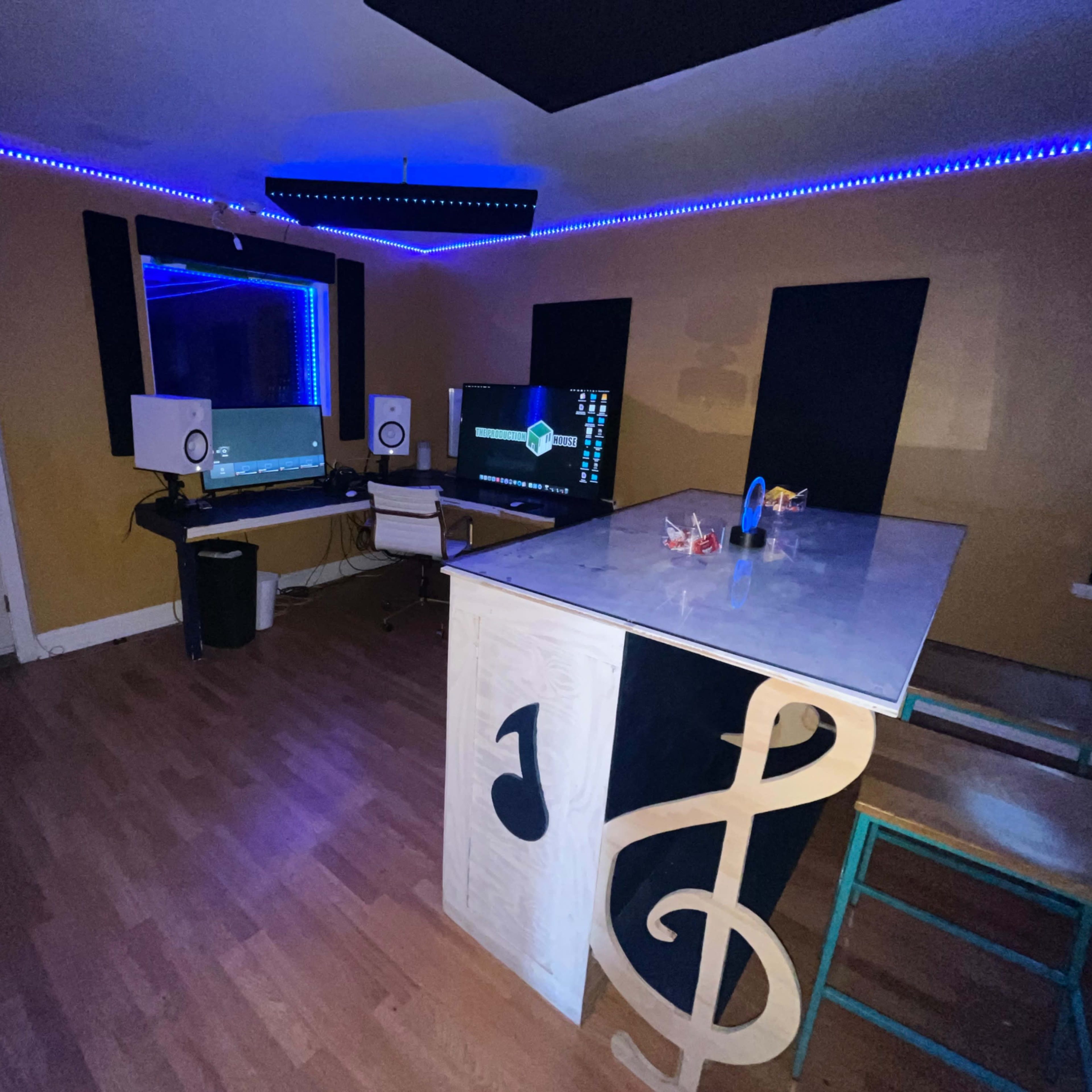 A small music studio featuring a desk with two monitors, studio speakers, and colorful LED strip lighting along the walls.