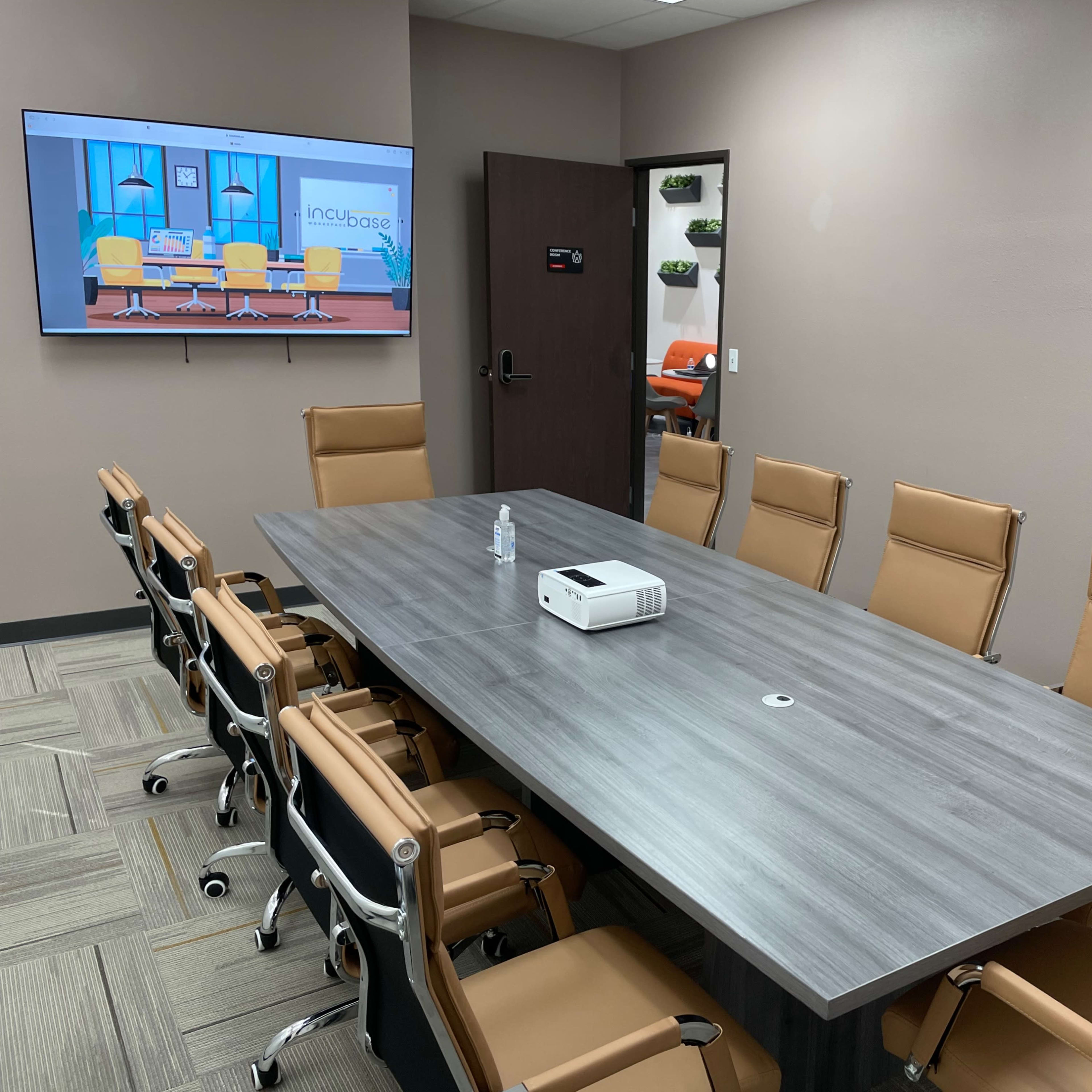 A conference room features a large gray table surrounded by multiple brown chairs, with a projector on the table and a screen displaying a colorful presentation on the wall.