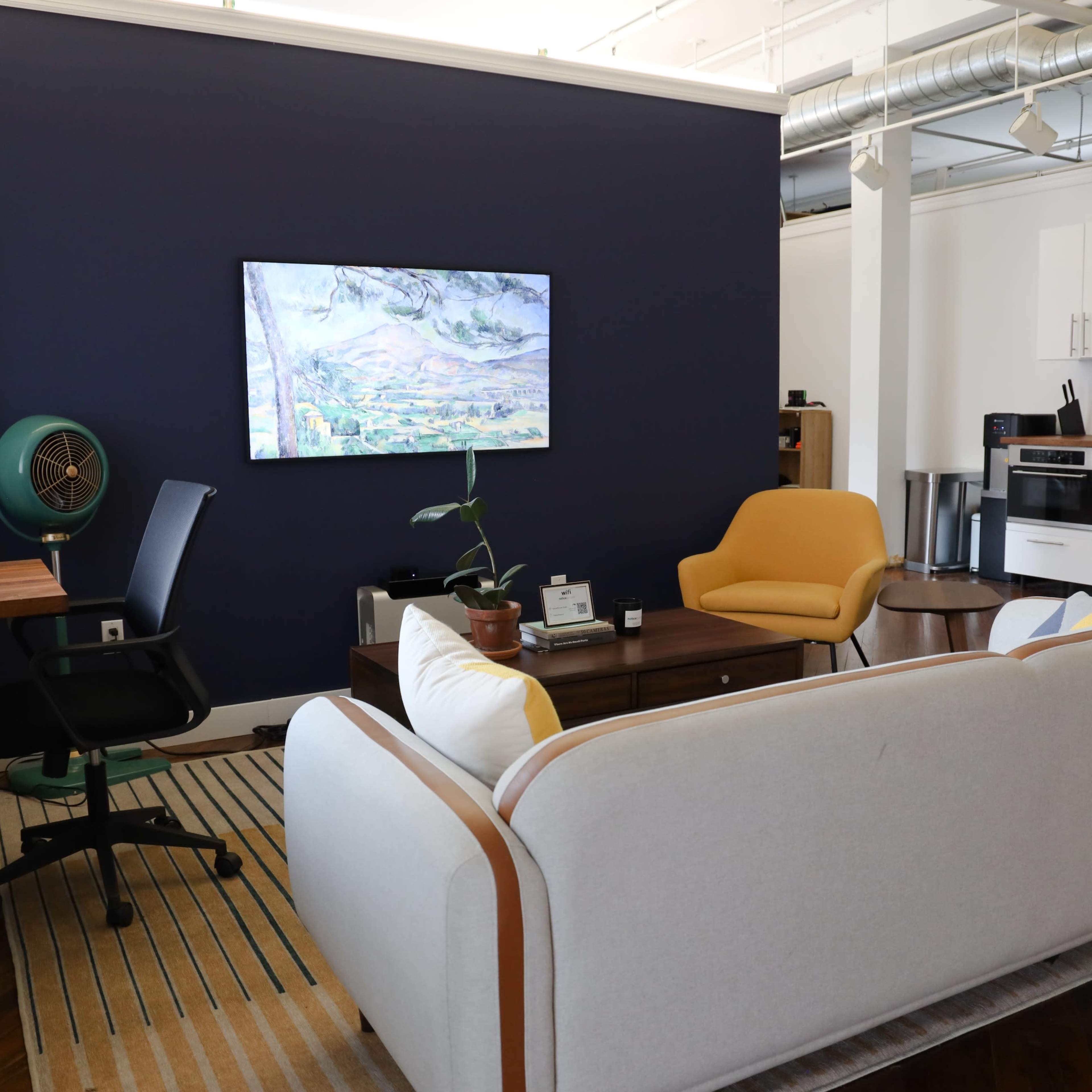 A modern living space featuring a light-colored sofa, a yellow chair, a television on the wall, and a kitchen area with white cabinets.