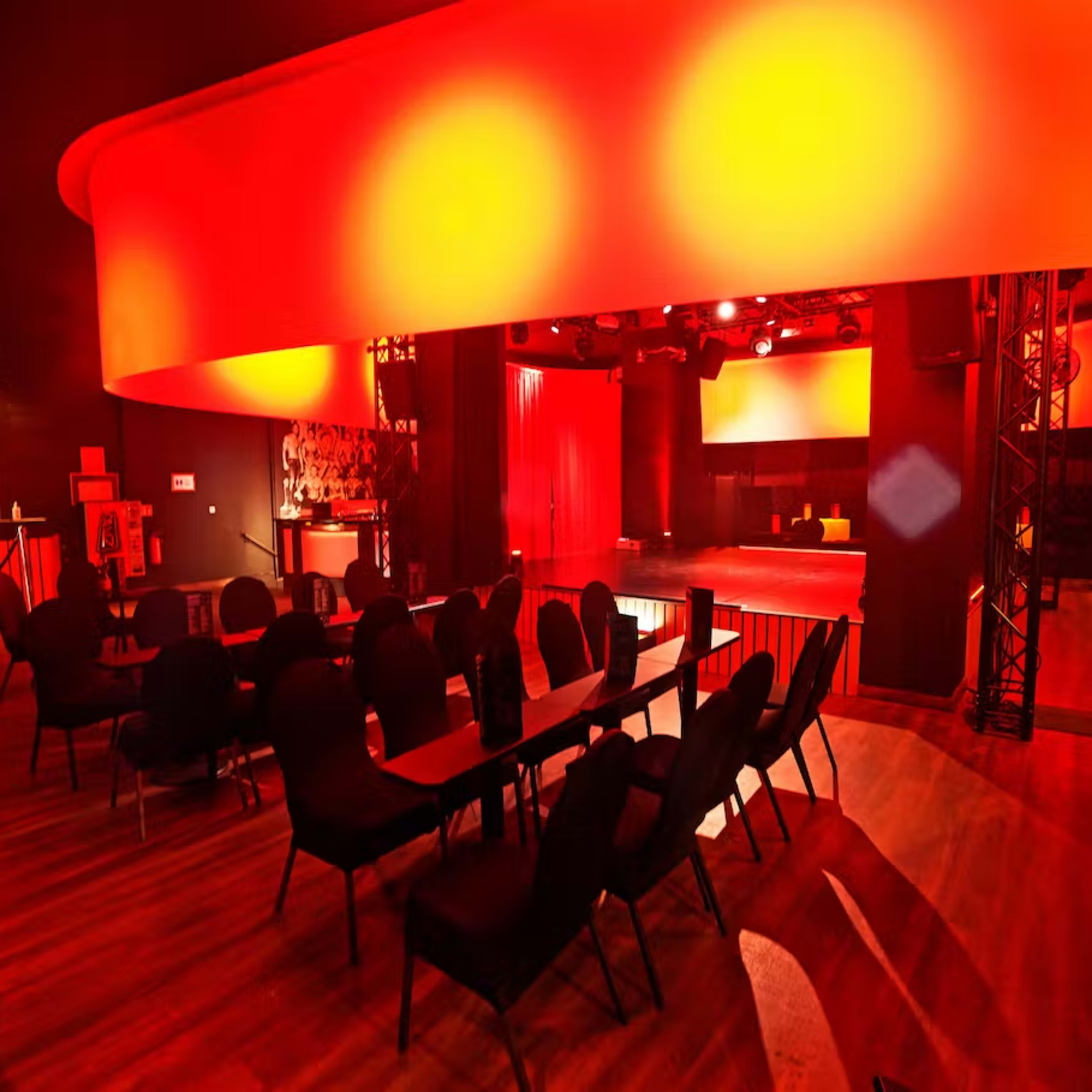 A dimly lit performance venue featuring a circular overhead light, rows of black chairs, and a stage area with red curtains.