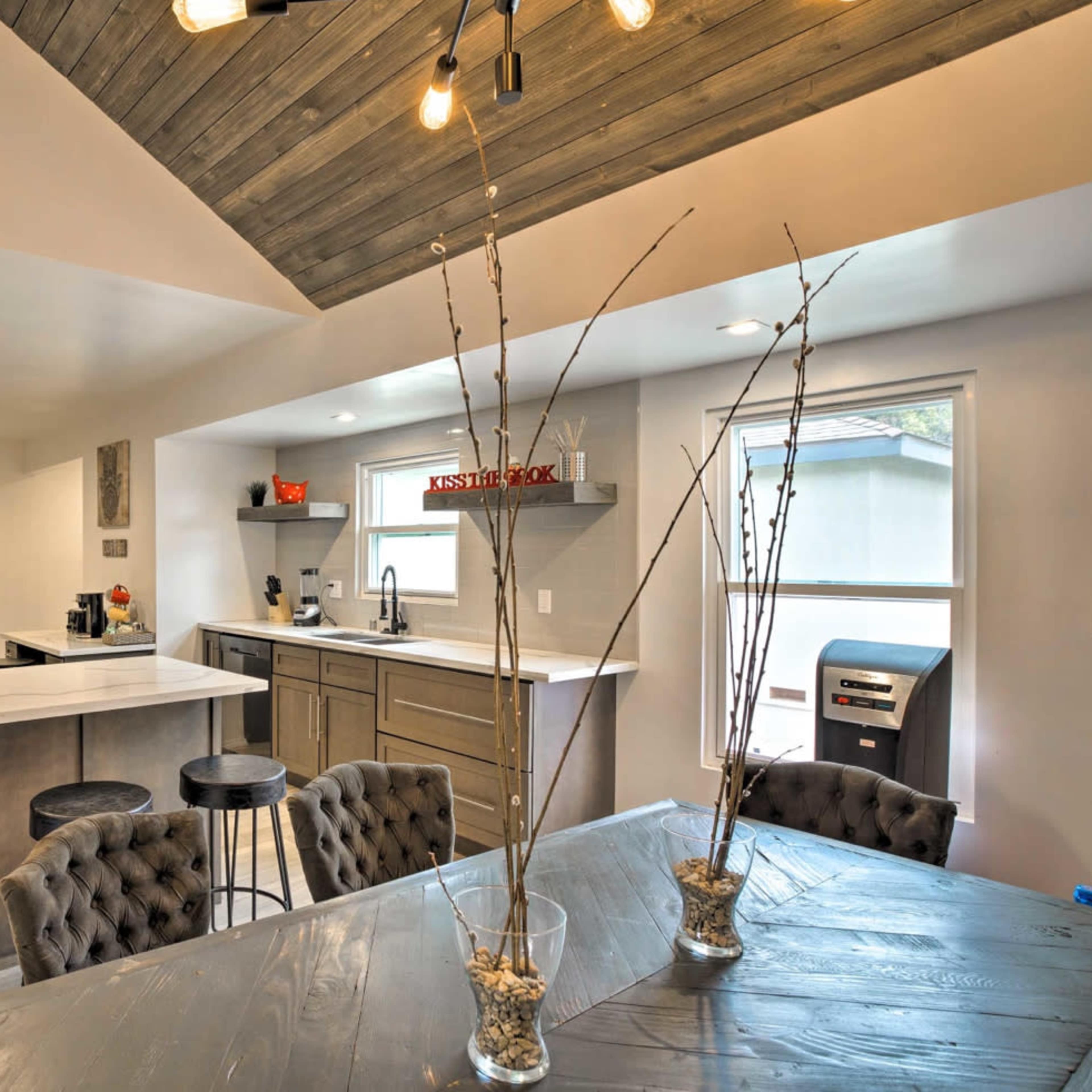 A modern kitchen and dining area featuring a white countertop, dark cabinetry, and a wooden ceiling with exposed lighting.