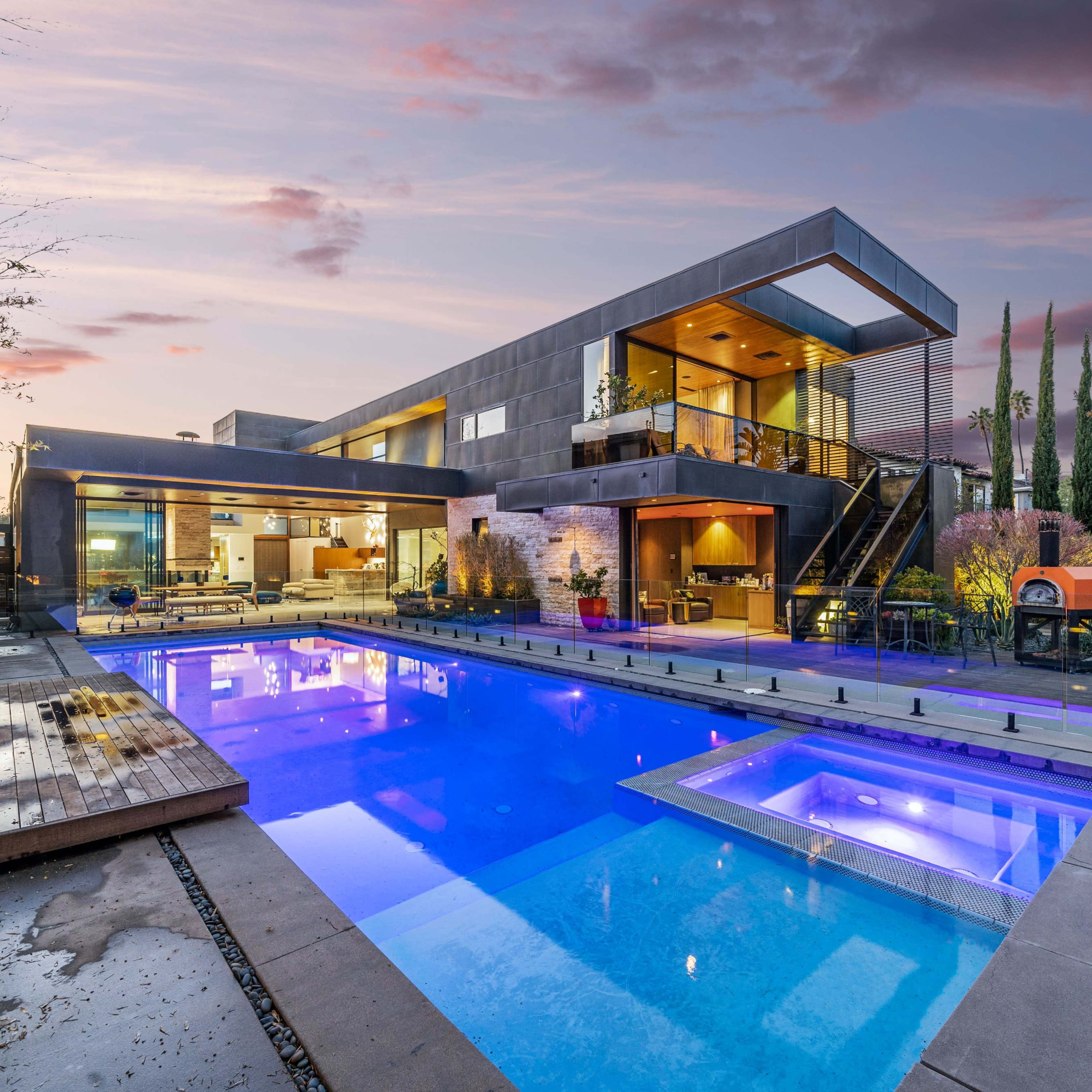 A modern two-story house with large windows and a prominent swimming pool in the foreground, surrounded by landscaped gardens.