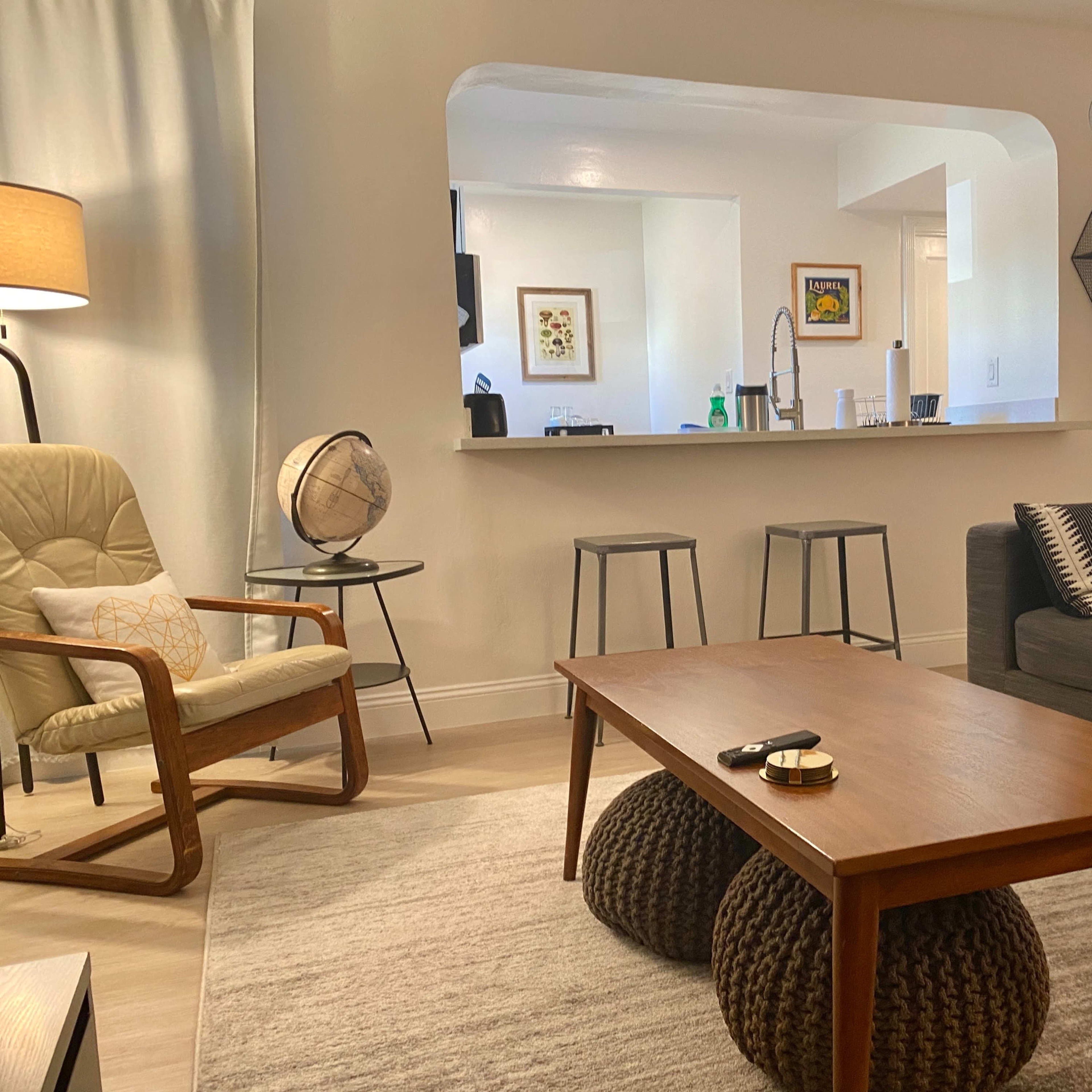 A modern living room with a wooden coffee table, a gray sofa, a beige armchair, and a kitchen area visible through an archway.