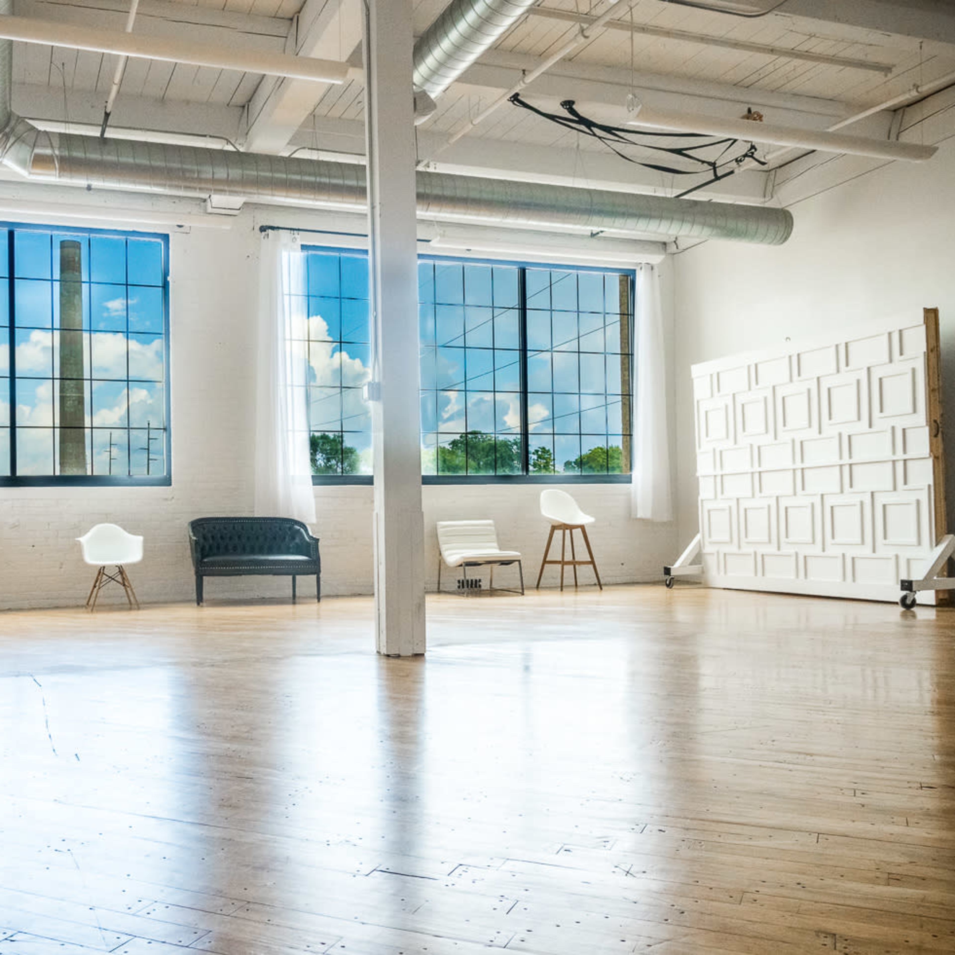 A spacious, bright room with wood flooring, large windows, and minimal furniture including a gray sofa and white chairs.