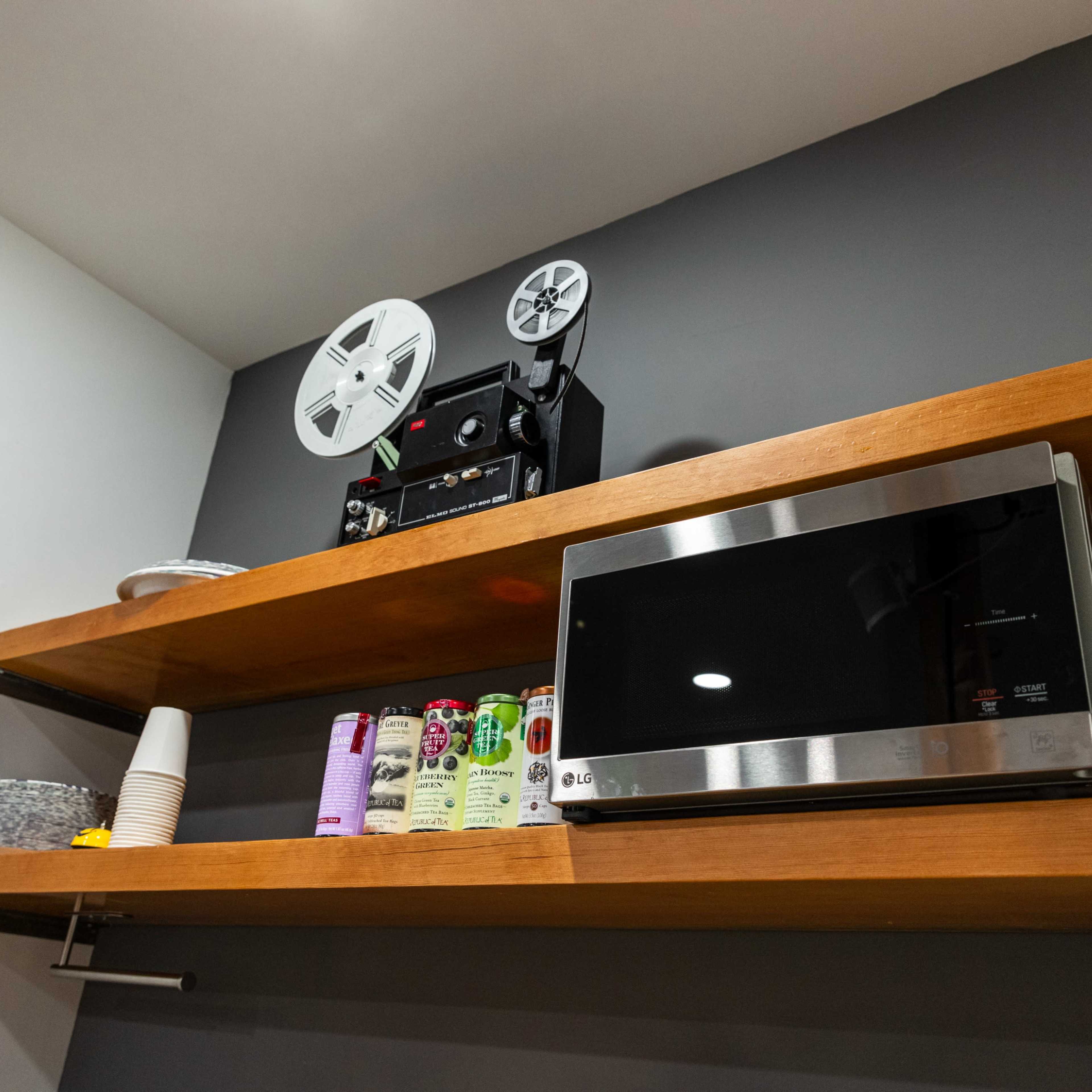 A black and silver microwave and a vintage film projector placed on wooden shelves against a dark grey wall.