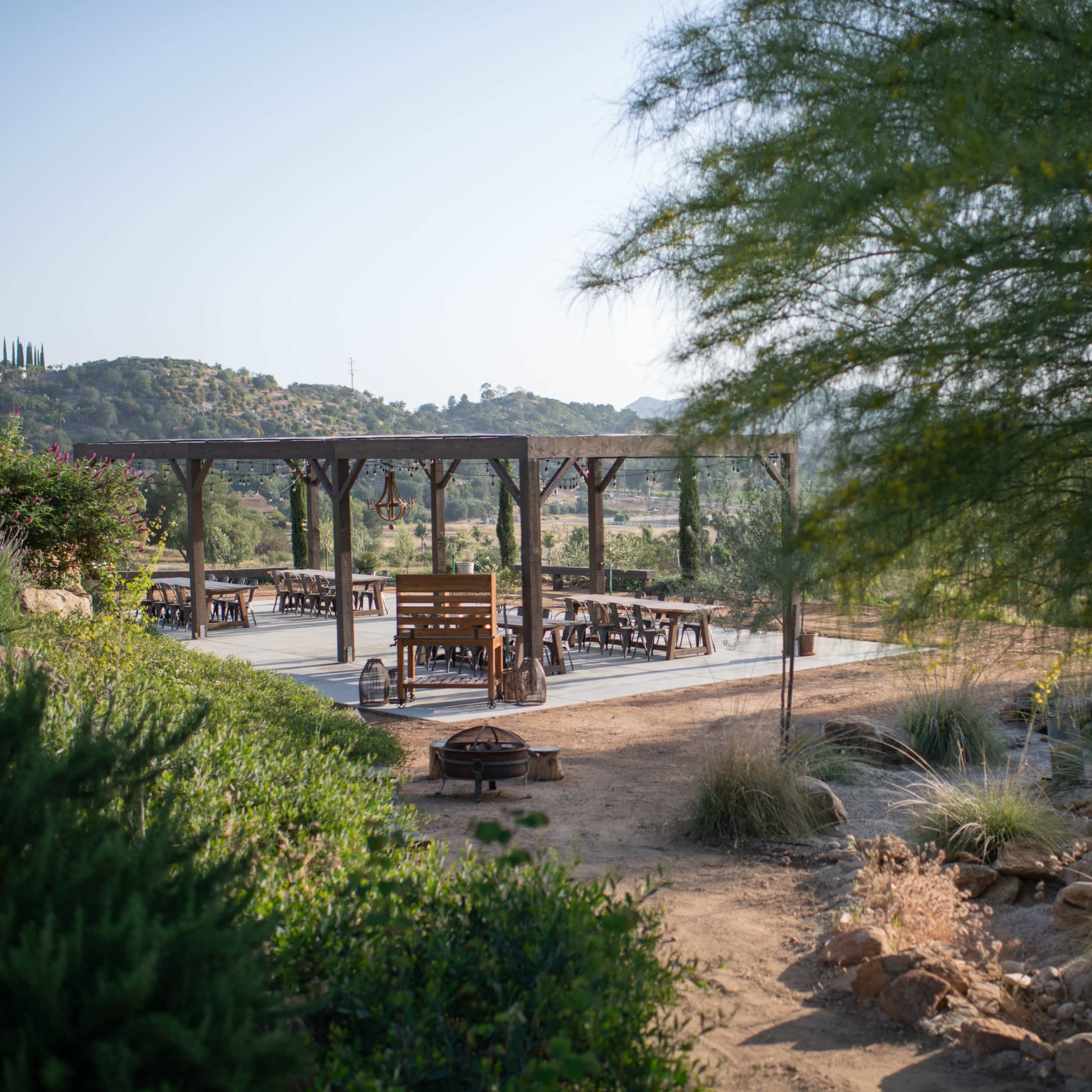 A wooden pavilion with seating surrounded by shrubs and mountains in the background.