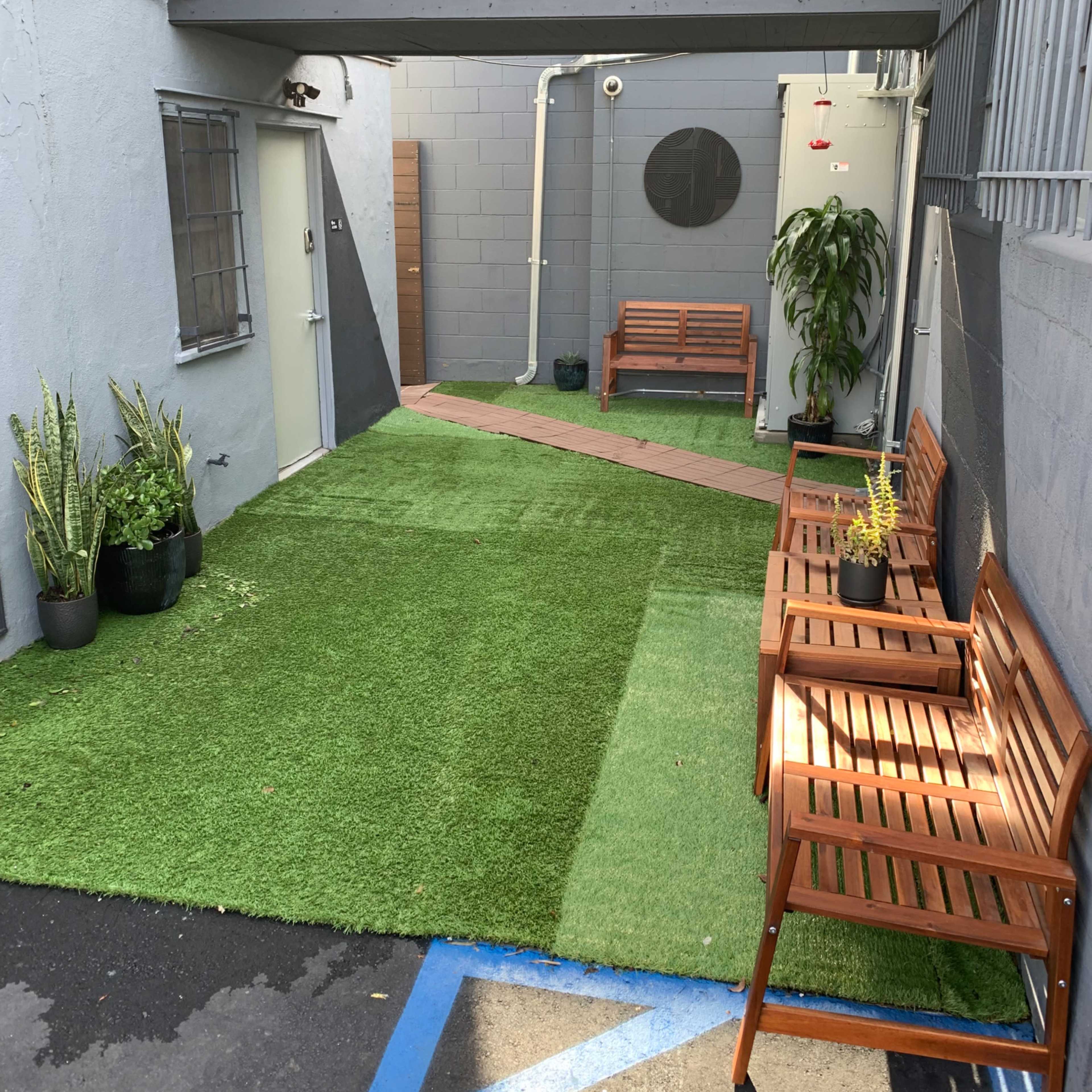 A narrow courtyard with artificial grass and wooden benches on either side, bordered by gray walls.